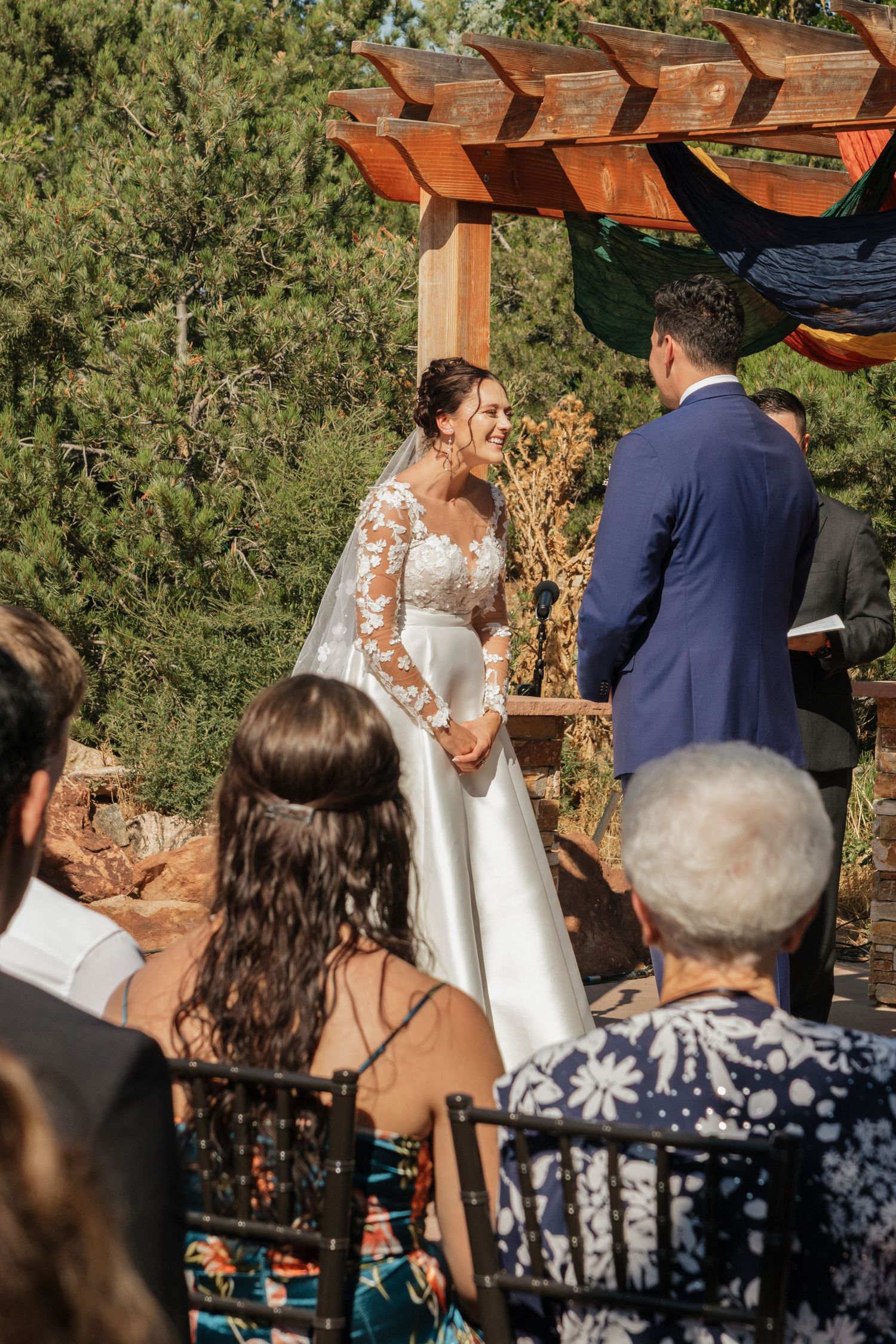 Wedding ceremony under wooden pergola with guests watching in foreground.