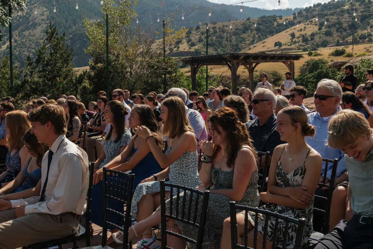 Wedding guests seated outdoors with mountains visible in the background on a sunny day.