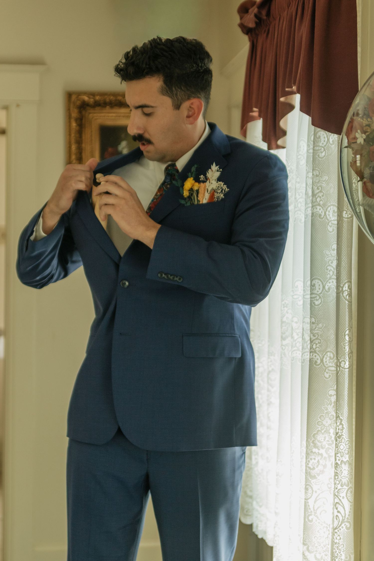 A person in a navy suit adjusts their tie while standing by lace curtains.