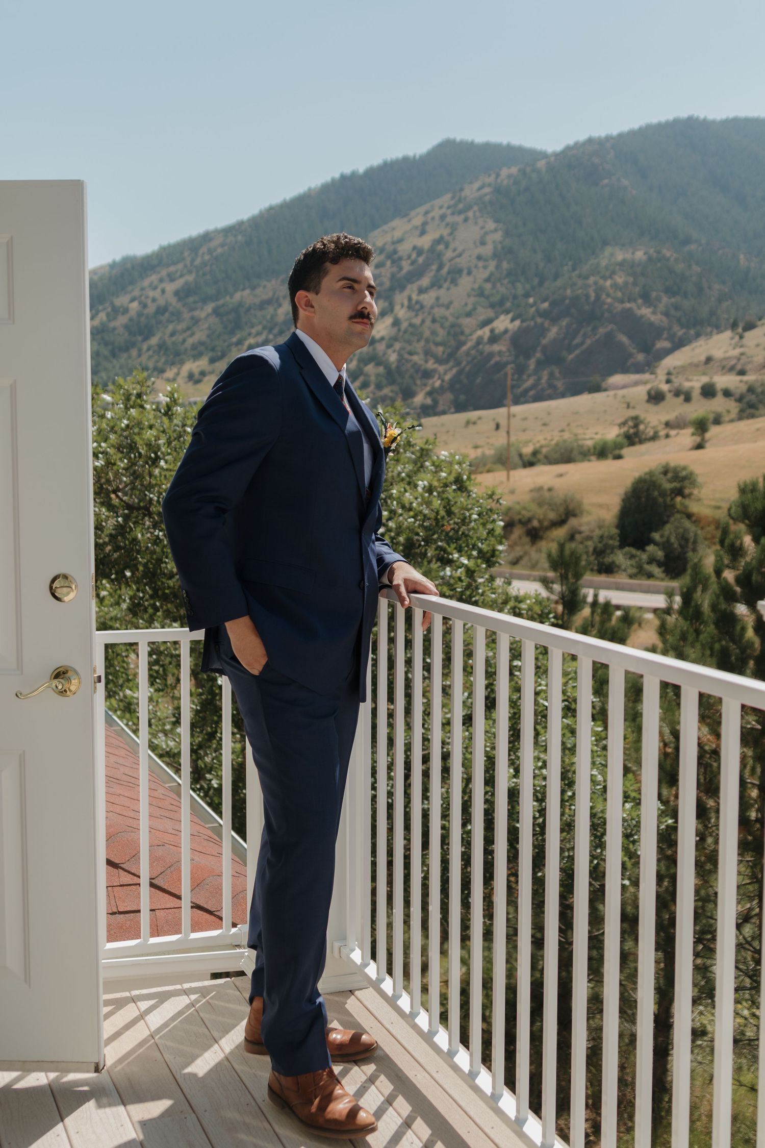 A person in a navy suit stands on a white railed porch overlooking mountain views.