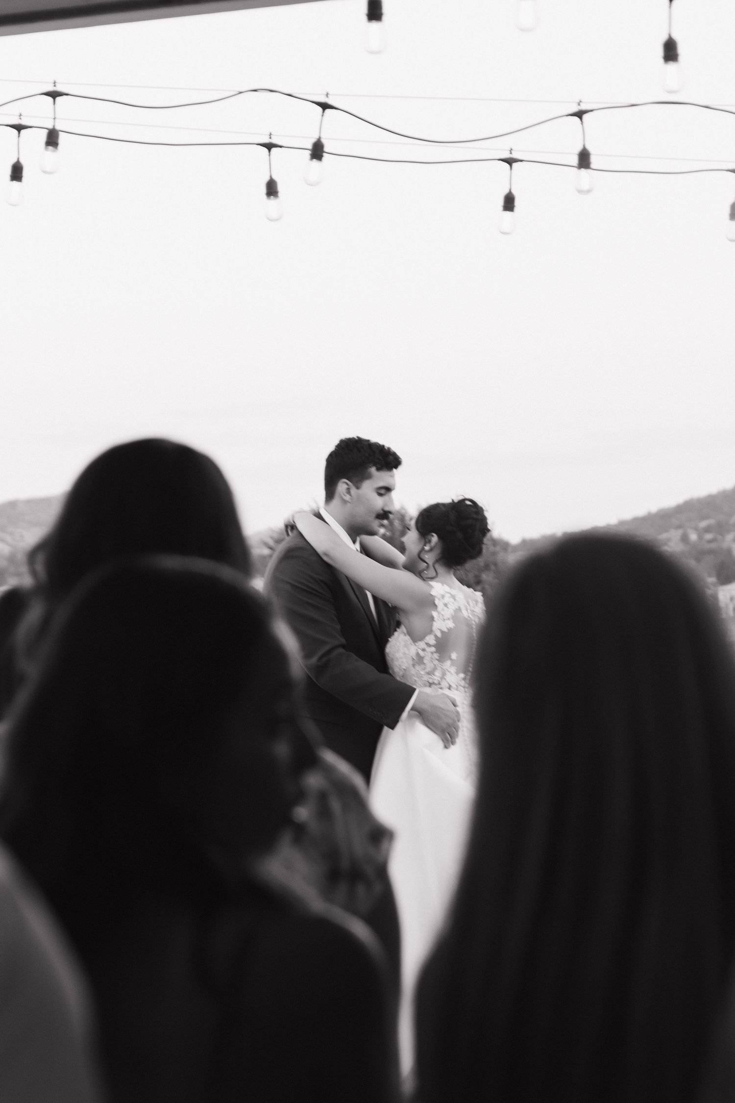 Black and white silhouette of wedding couple under string lights at dusk.