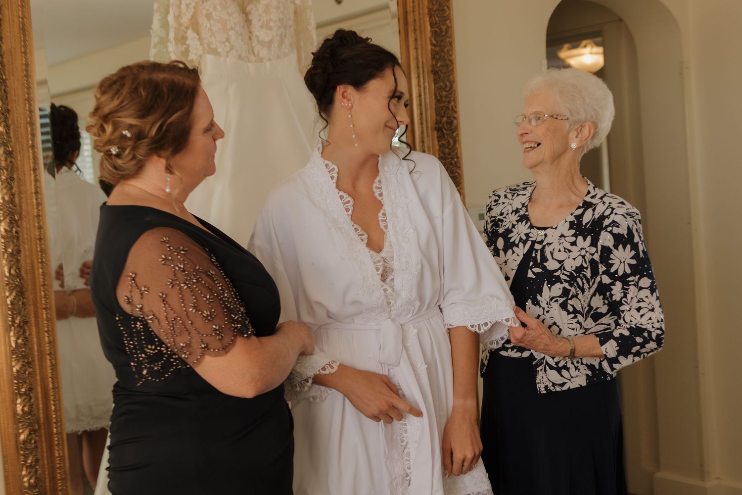 Three individuals share a joyful moment while getting ready in elegant black and white attire for a special occasion.