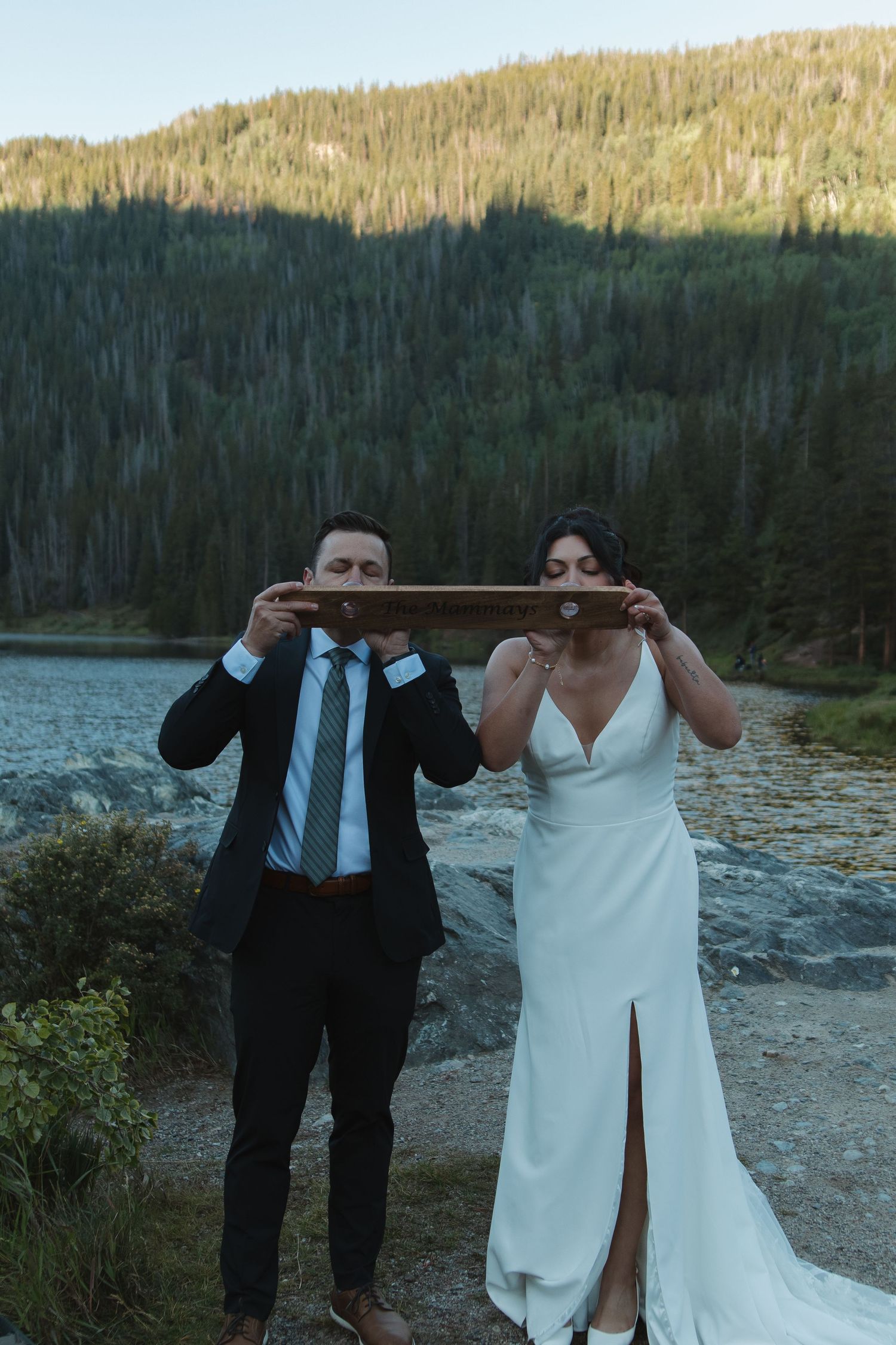 A couple in wedding attire shares playful moments outdoors by a mountain lake.