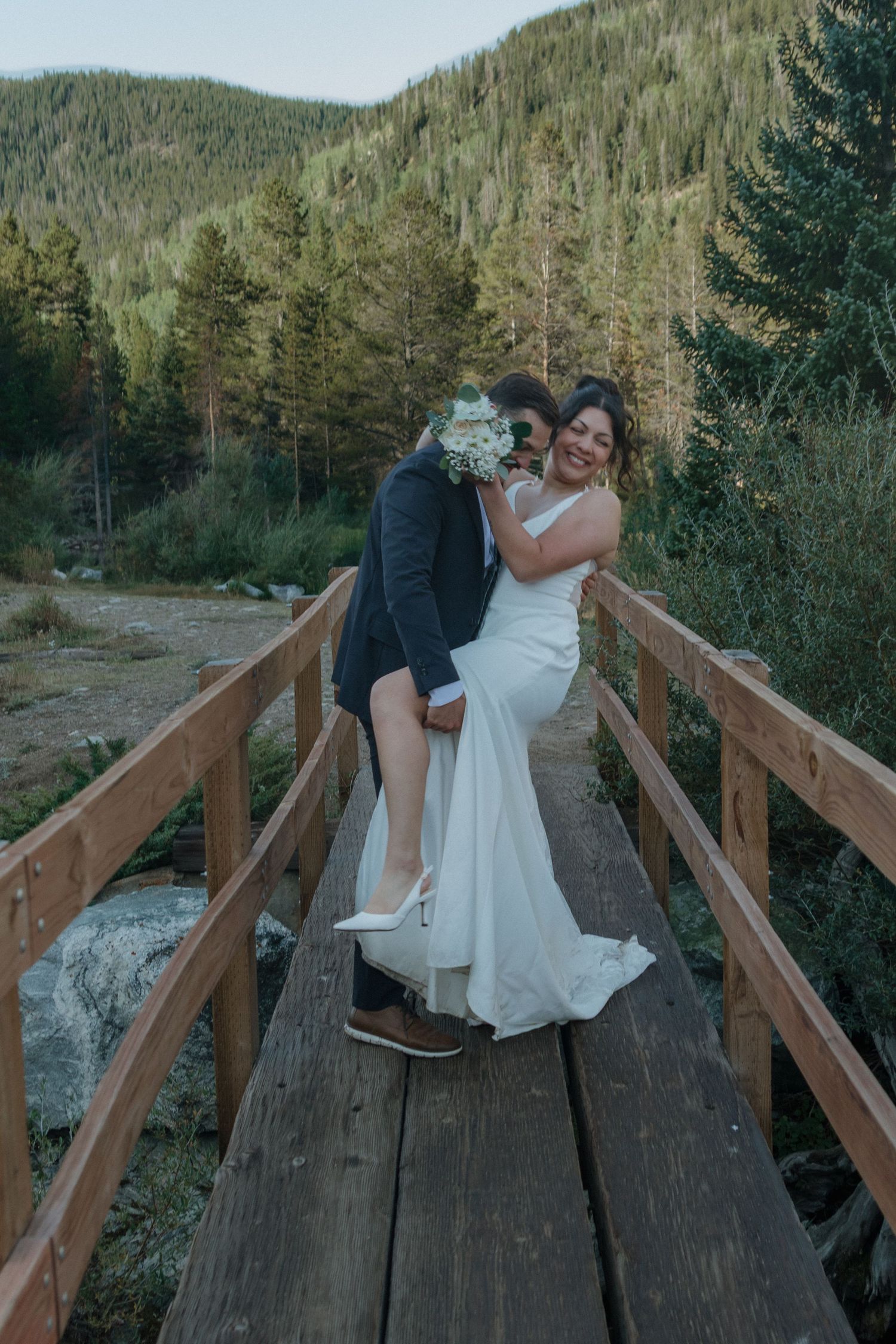 A romantic moment captured on a wooden footbridge surrounded by mountain scenery.