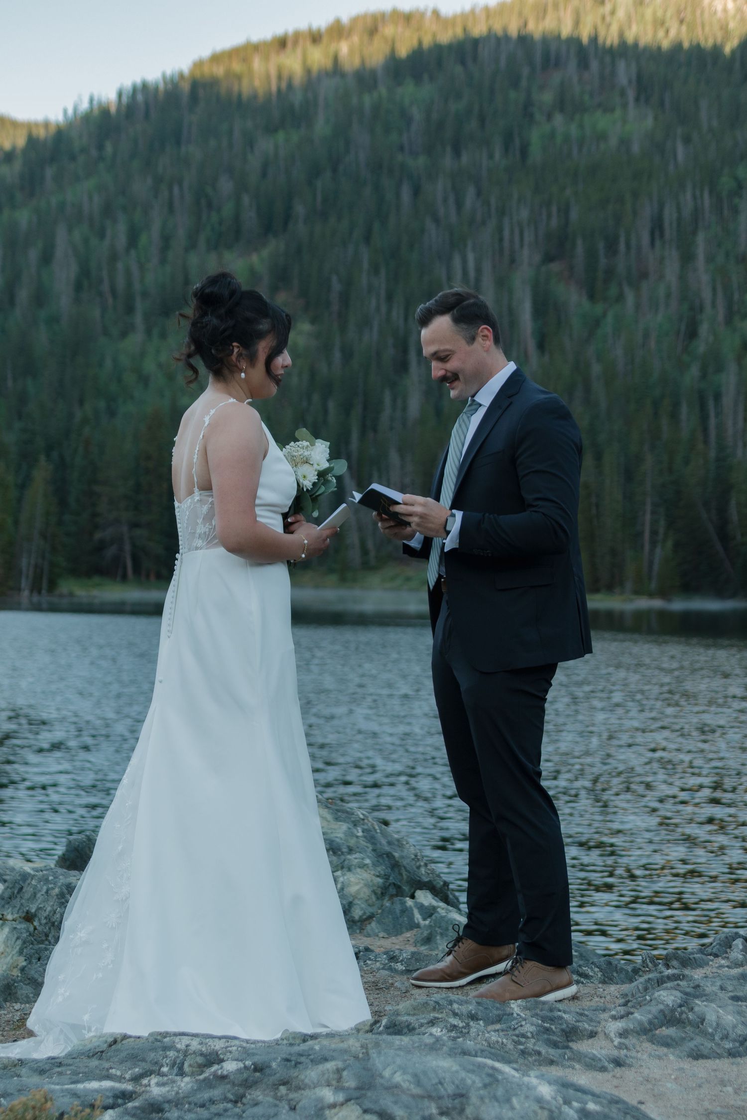 A romantic lakeside wedding exchange of vows on a beach with mountain and forest scenery in the background.