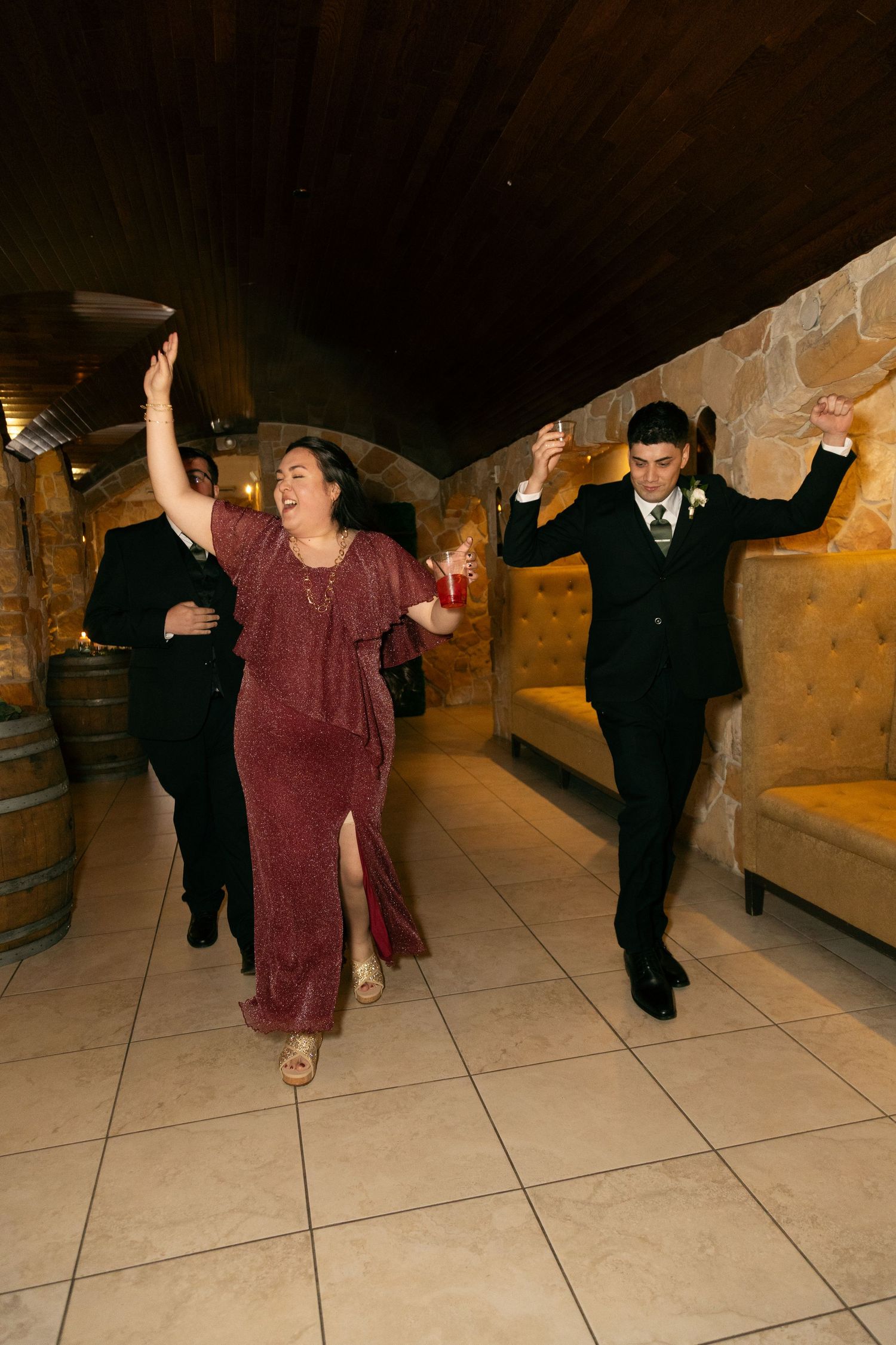 Woman in burgundy dress and groom in suit dance energetically at indoor wedding reception.