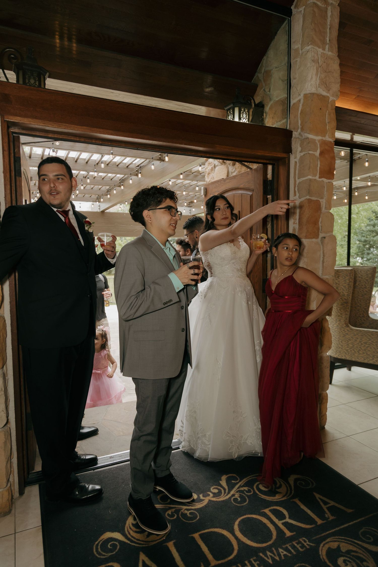 Bride in white lace gown with wedding guests at venue entrance pointing toward the reception area.