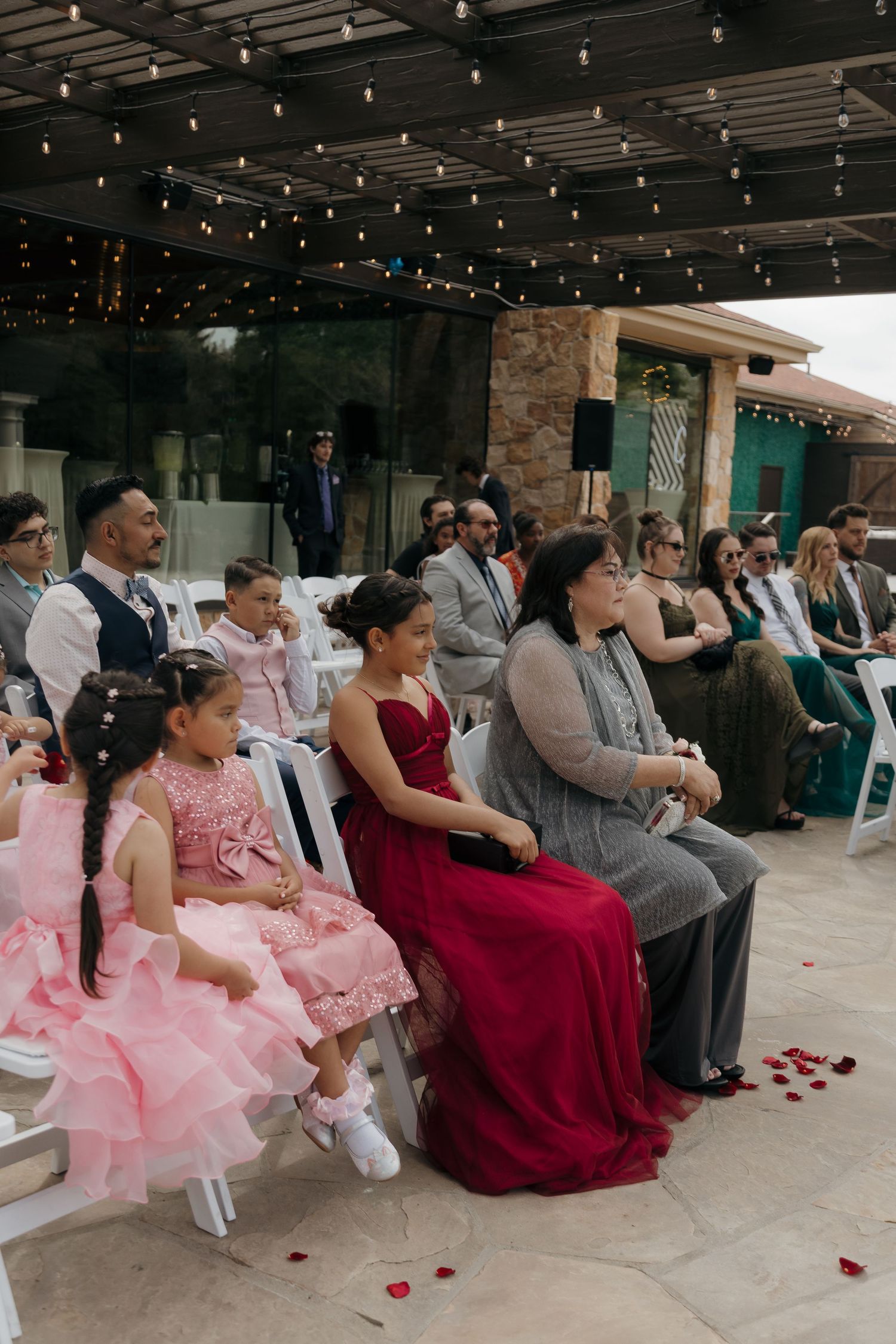 Wedding guests seated outdoors under string lights watching ceremony, including children in pink dresses and woman in red gown.