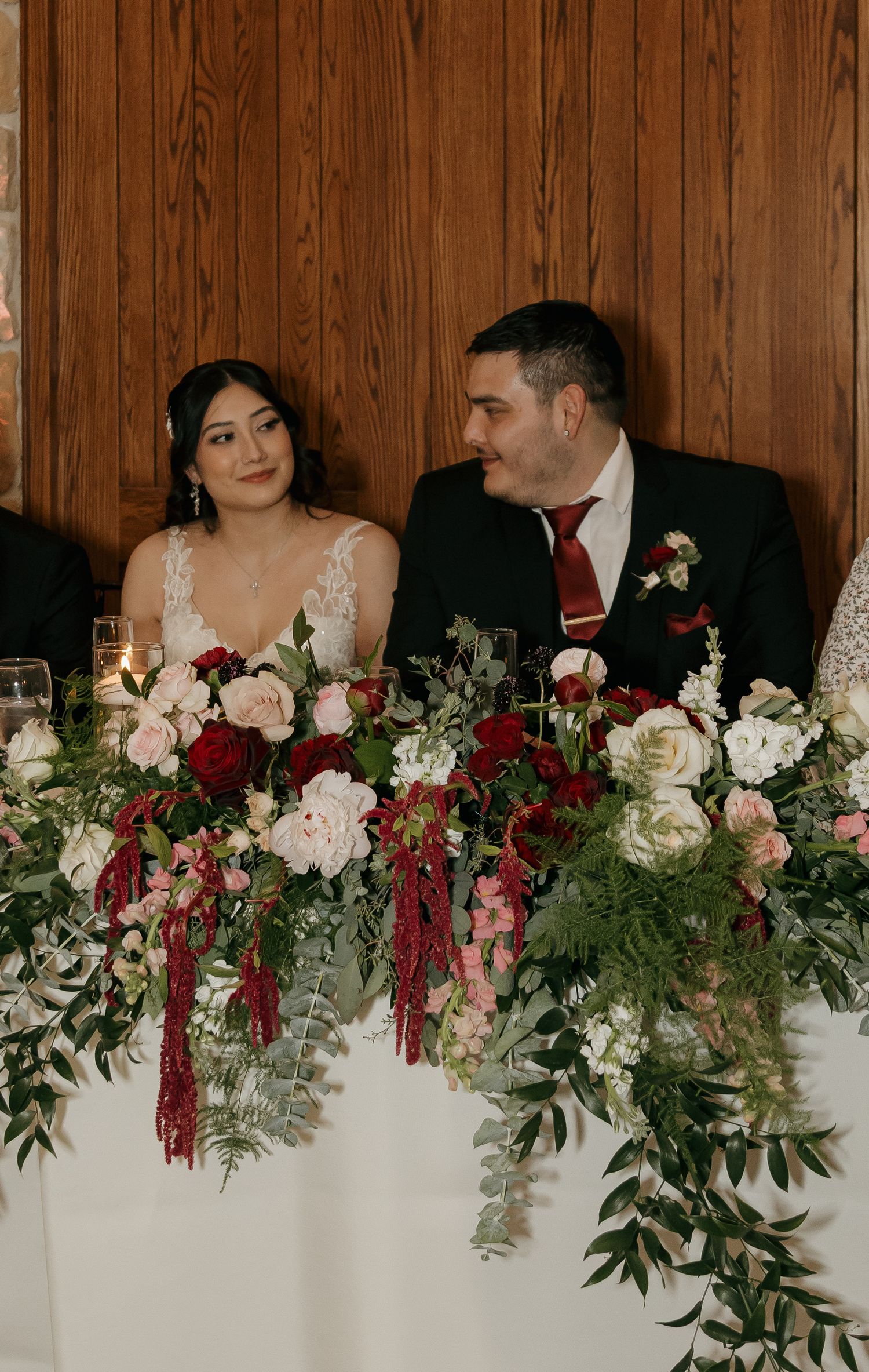 Bride and groom smile at each other at sweetheart table with stunning red and pink floral arrangements.