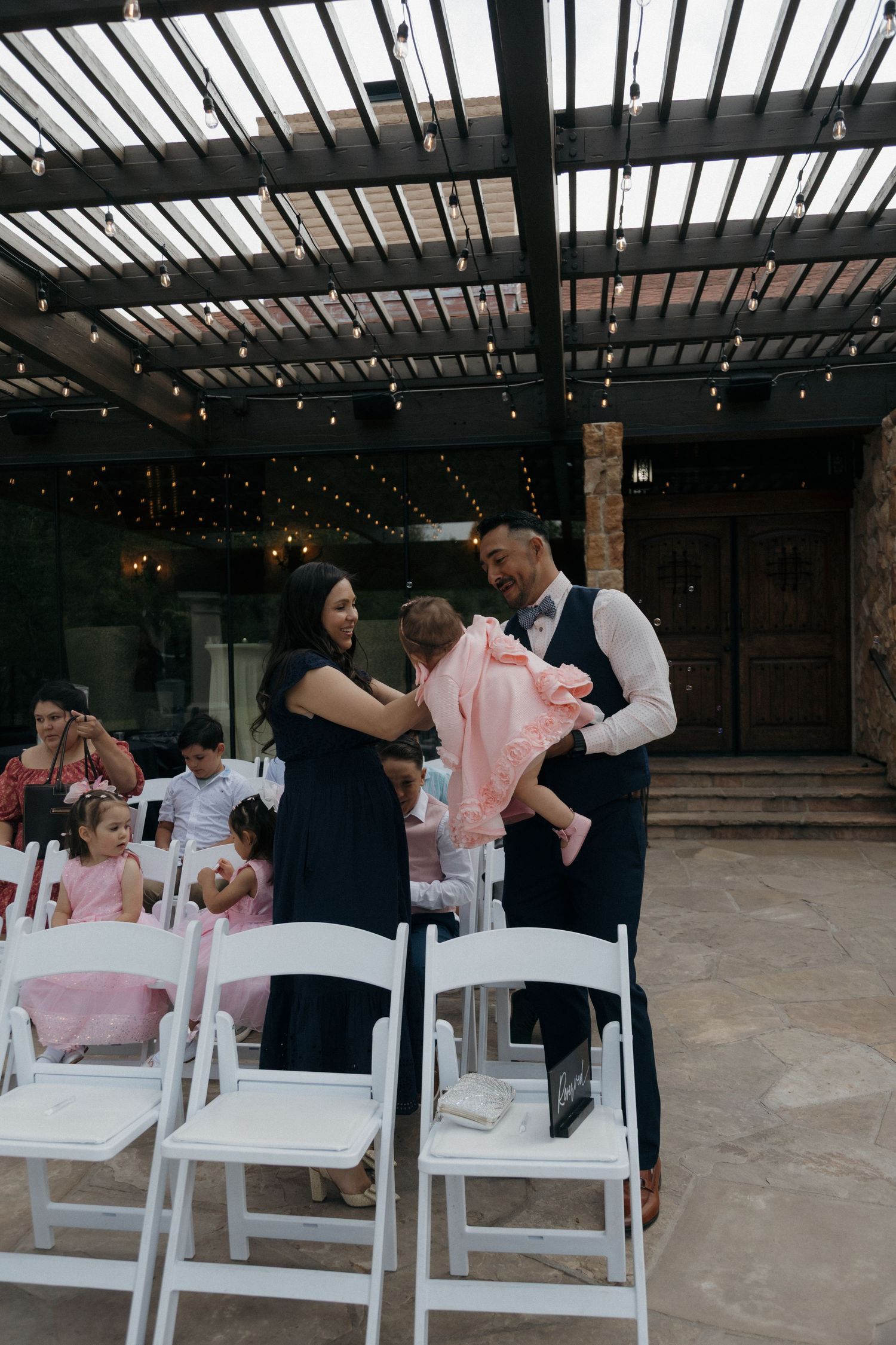 Man and woman interacting with a baby in pink outfit during an outdoor wedding ceremony with white chairs.