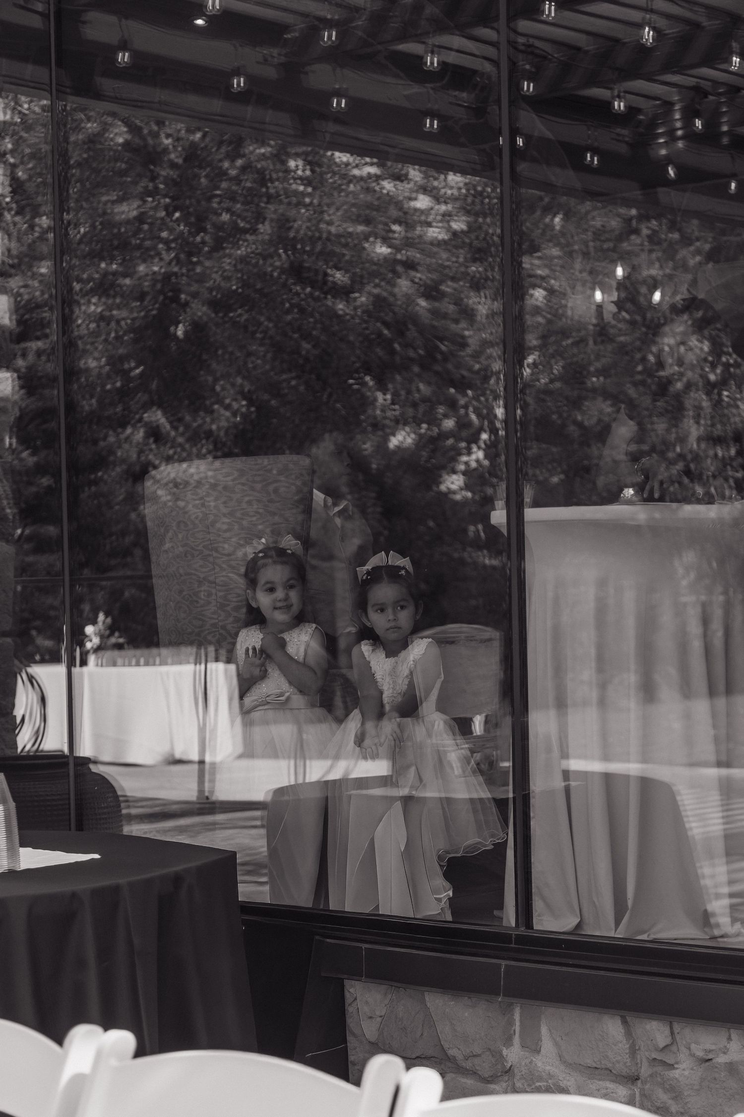 Black and white photo of two young flower girls in white dresses reflected in glass windows at wedding.