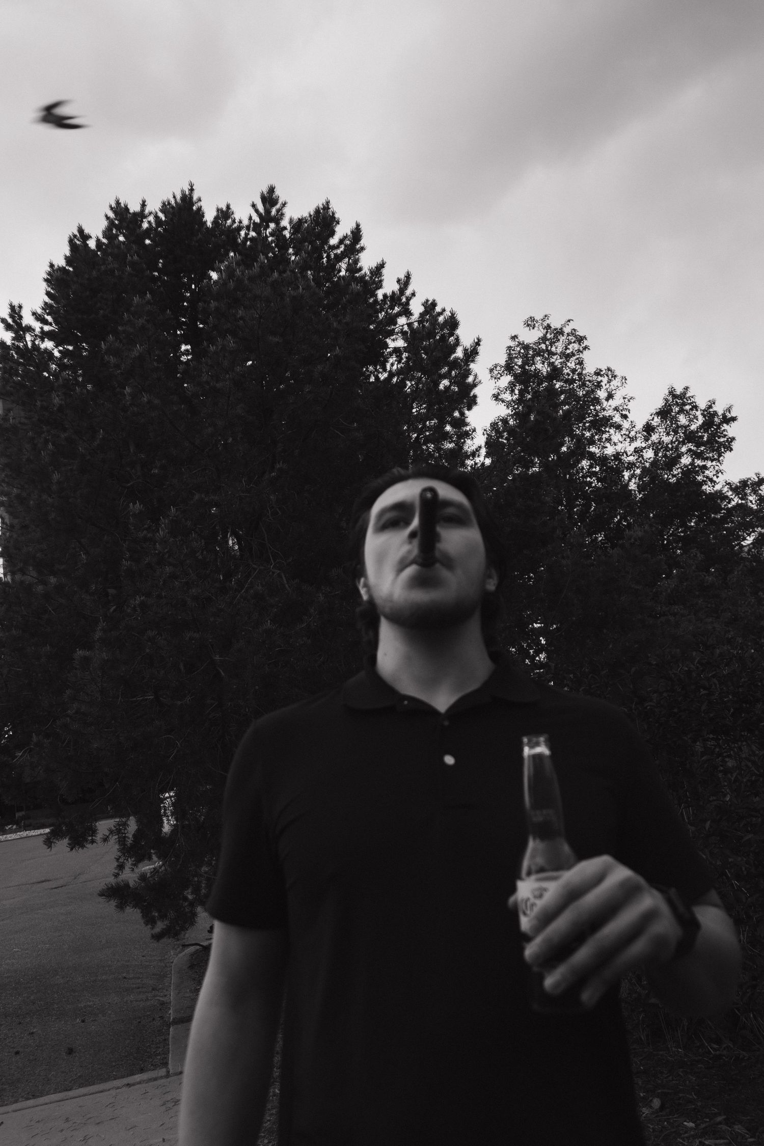 Black and white portrait of a young man holding a beer bottle outdoors with trees and cloudy sky behind him.
