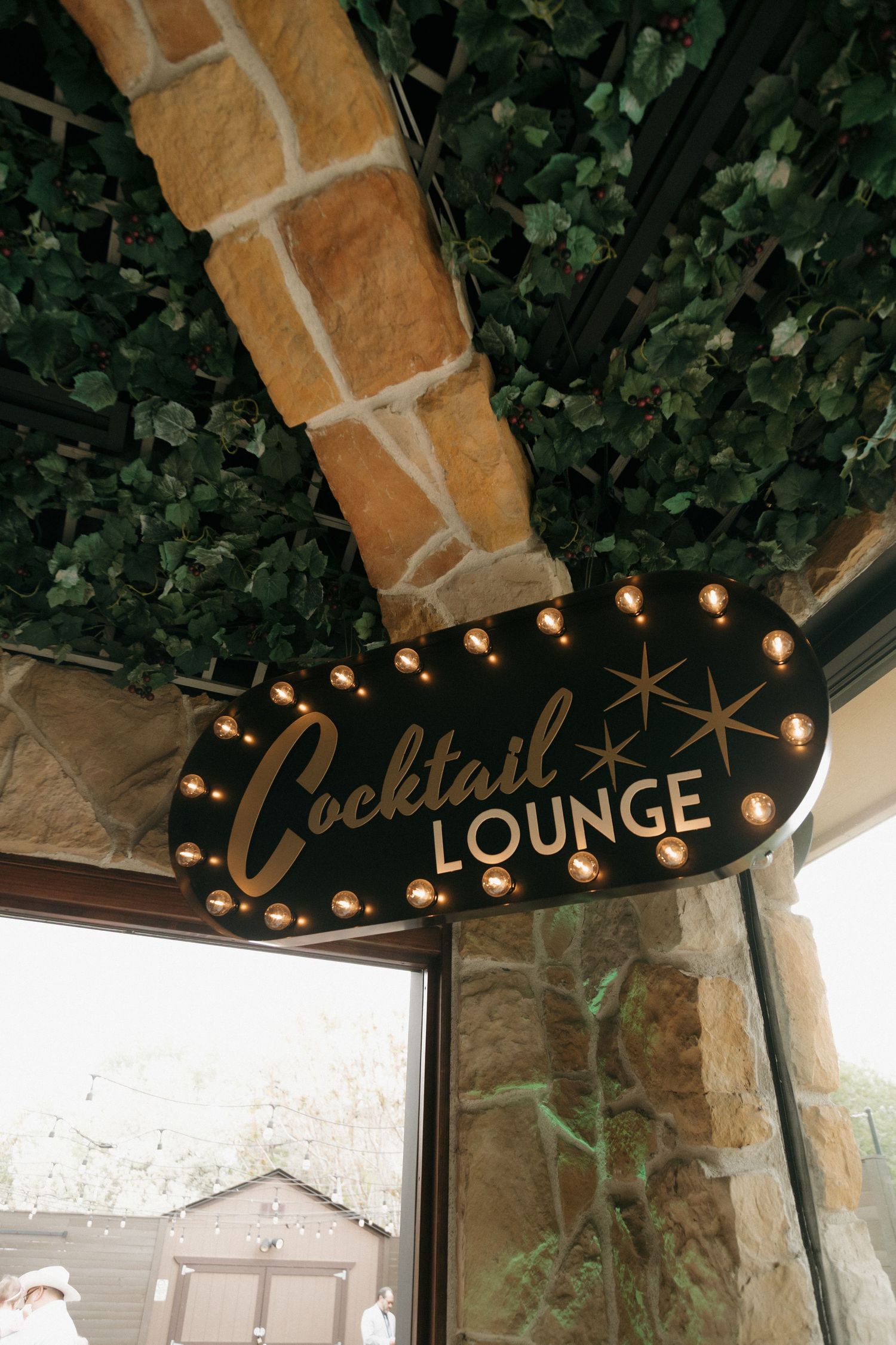 Illuminated Cocktail Lounge marquee sign with Edison bulbs mounted on stone wall with lush green ivy ceiling.