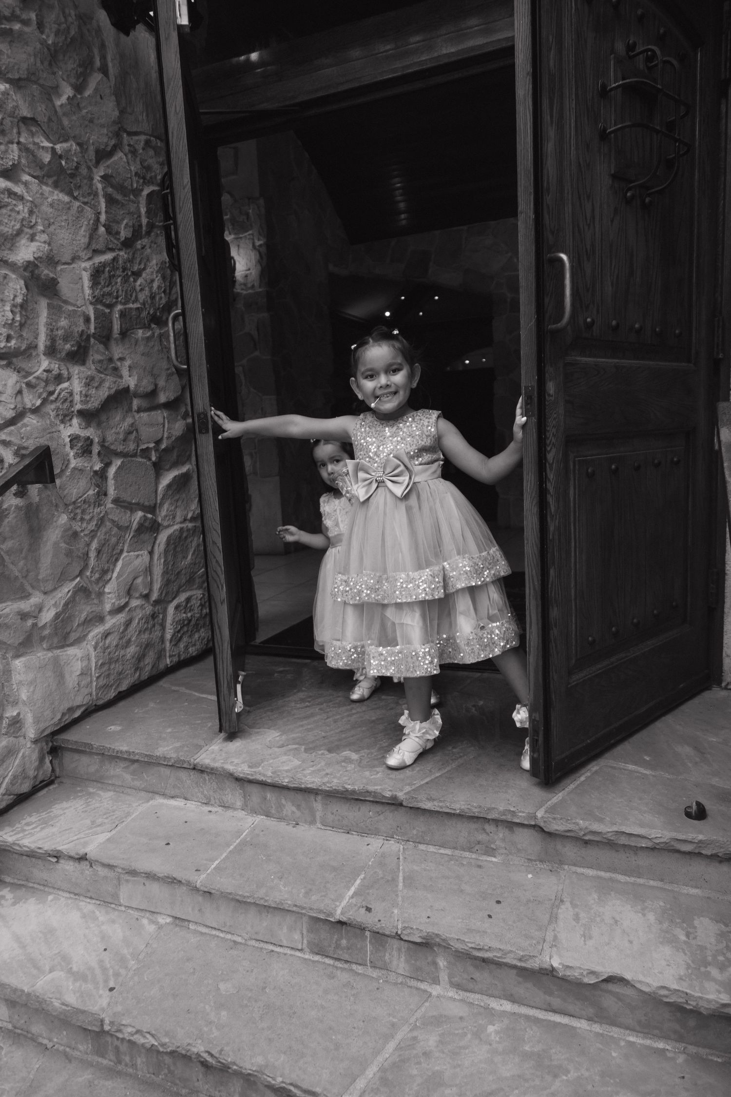 Black and white photo of young girl in sparkly dress smiling at open wooden church doors.