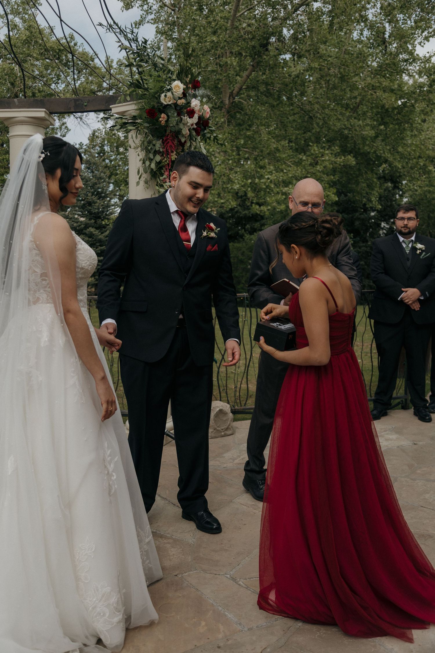 Bride and groom share a joyful moment at outdoor wedding ceremony with bridesmaid in burgundy dress.