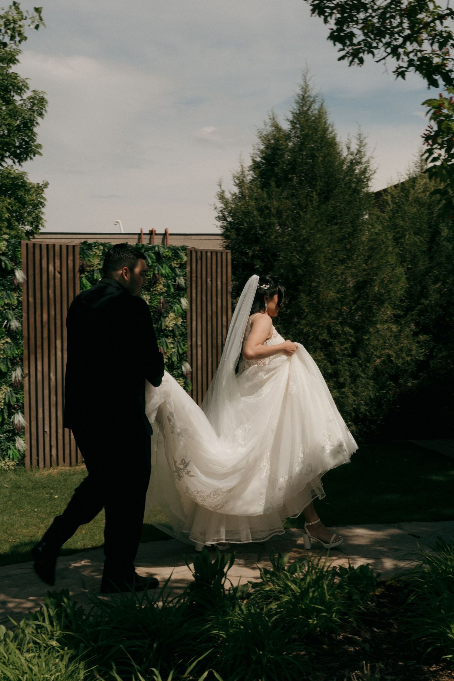 Groom in black suit carries bride's white lace wedding gown train as couple walks through garden.