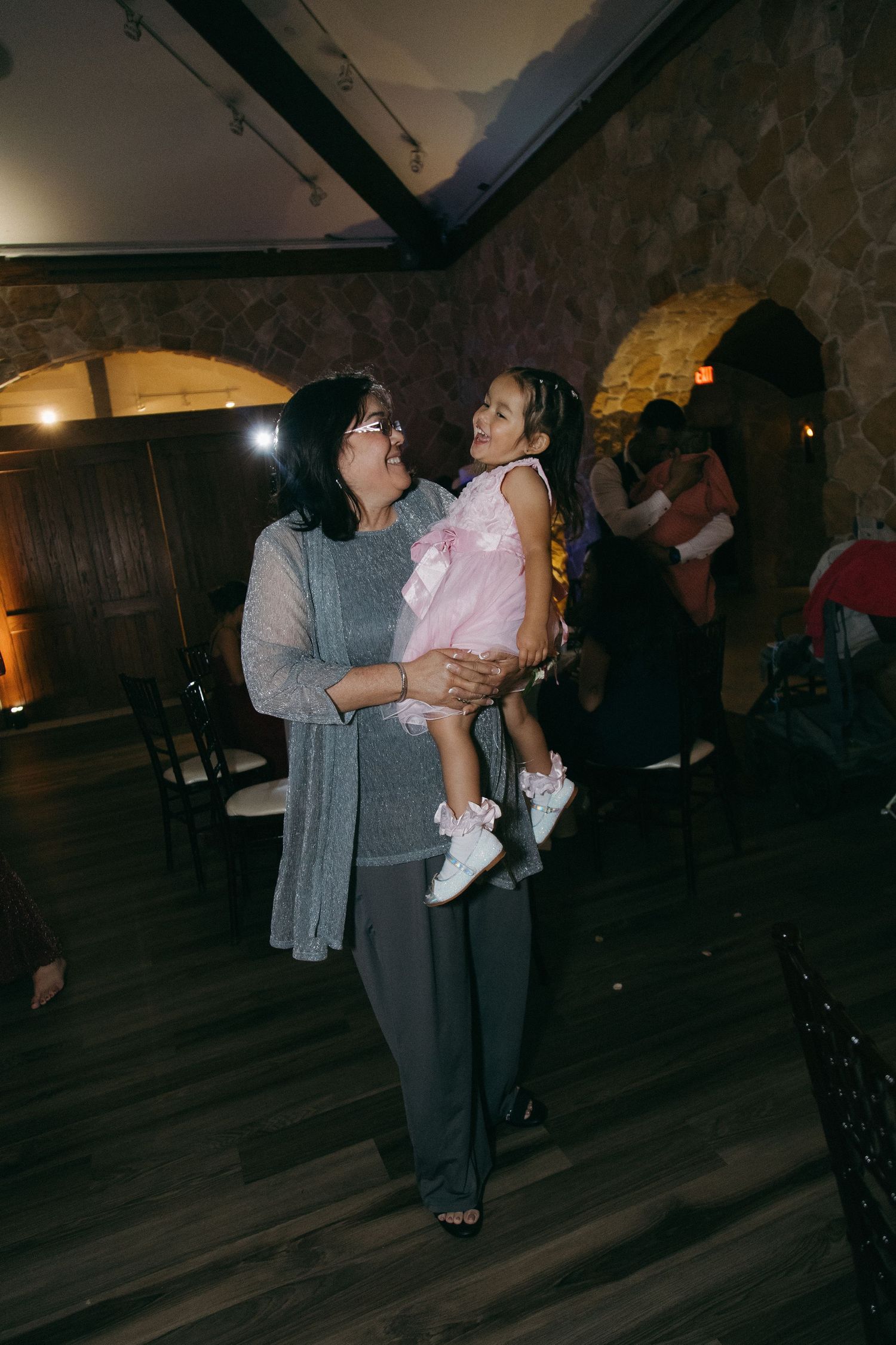 Grandmother holds young girl in pink dress while dancing together at a warmly lit wedding reception.