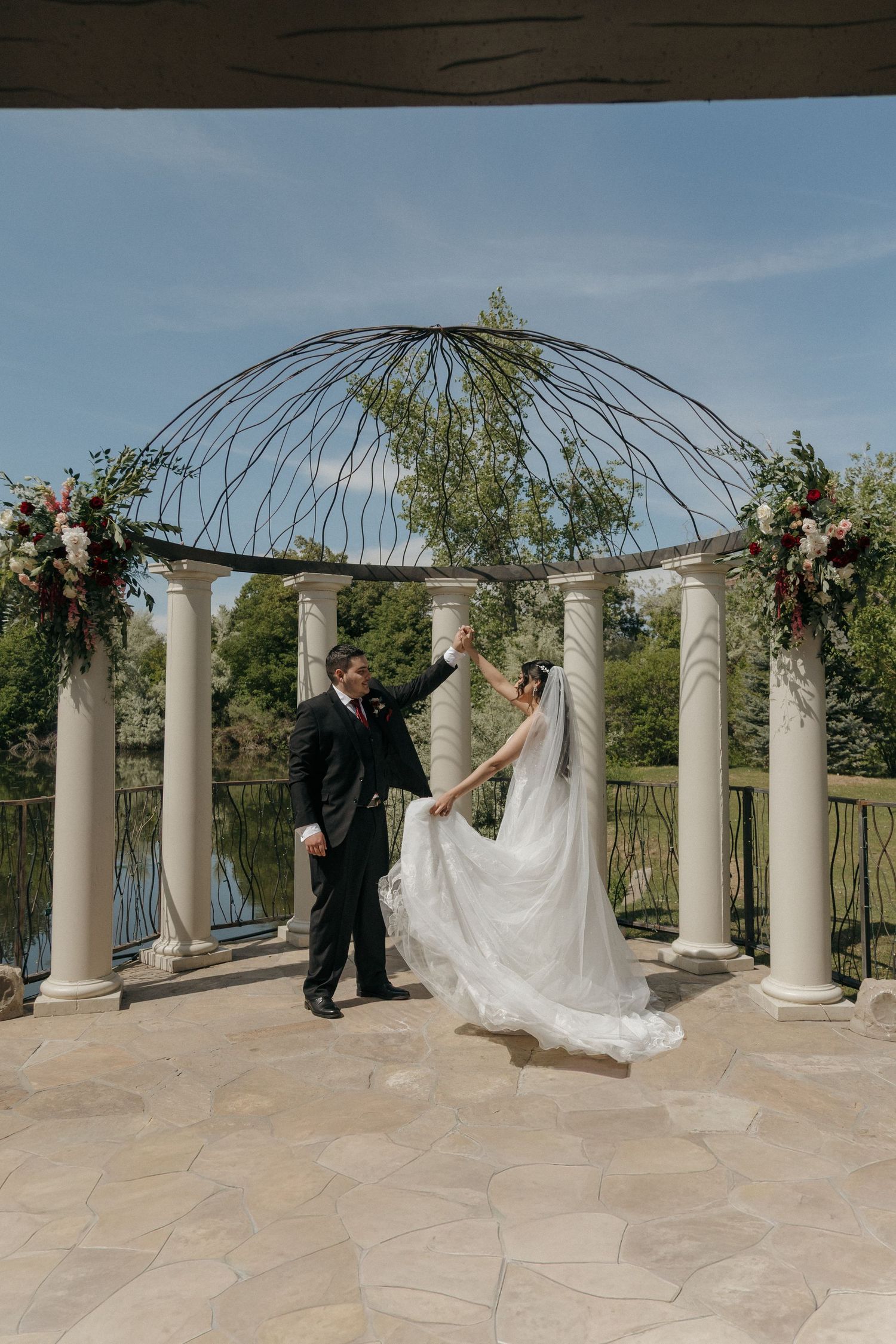 Bride and groom dancing under an elegant floral gazebo with a serene pond backdrop on their wedding day.