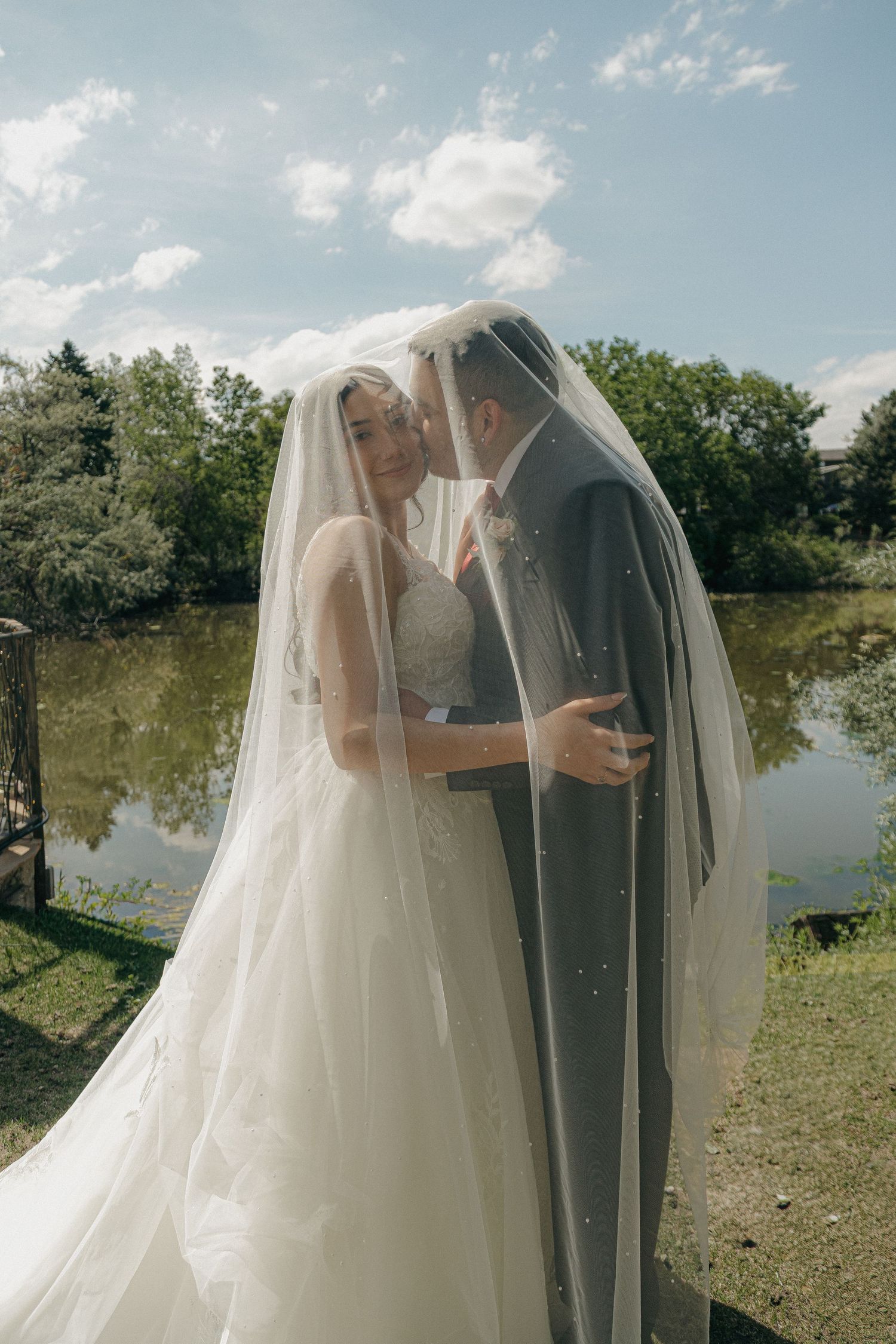 Bride and groom sharing a romantic kiss under a flowing veil beside a serene lake.
