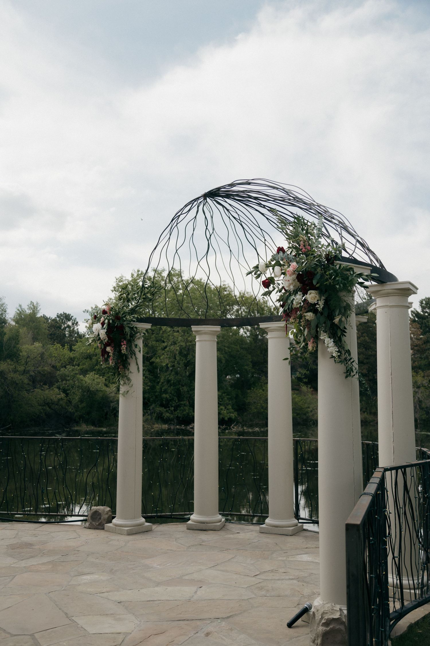 Elegant outdoor wedding arch with white columns, floral arrangements, and decorative branches by a lake.