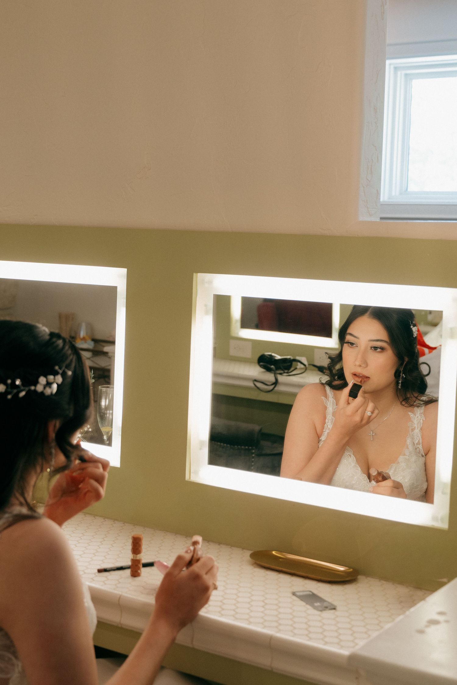 Bride applying lipstick at a lighted vanity mirror while wearing a floral hair piece and lace wedding dress.