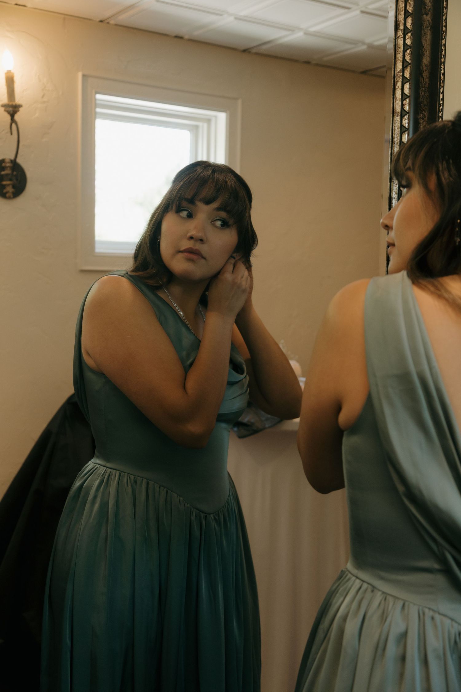 Bridesmaid in teal satin gown adjusting earrings while looking in a mirror inside a wedding venue suite.