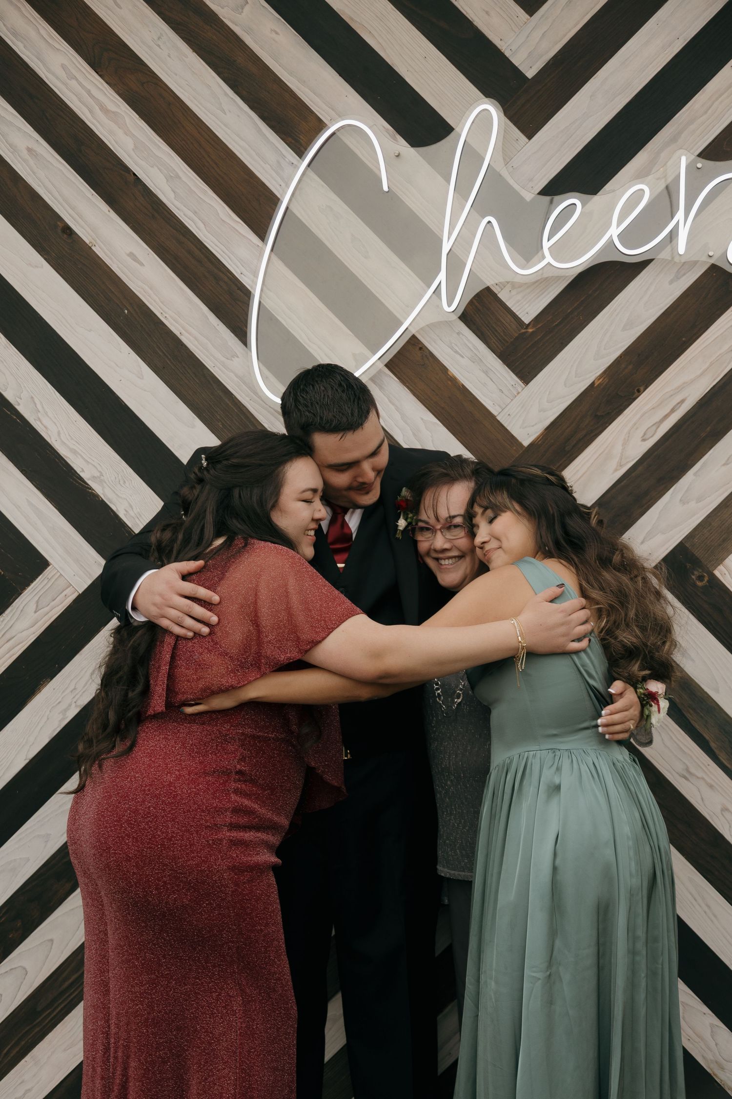 Groom and three women share a group hug in front of a chevron wood wall with neon Cheers sign.