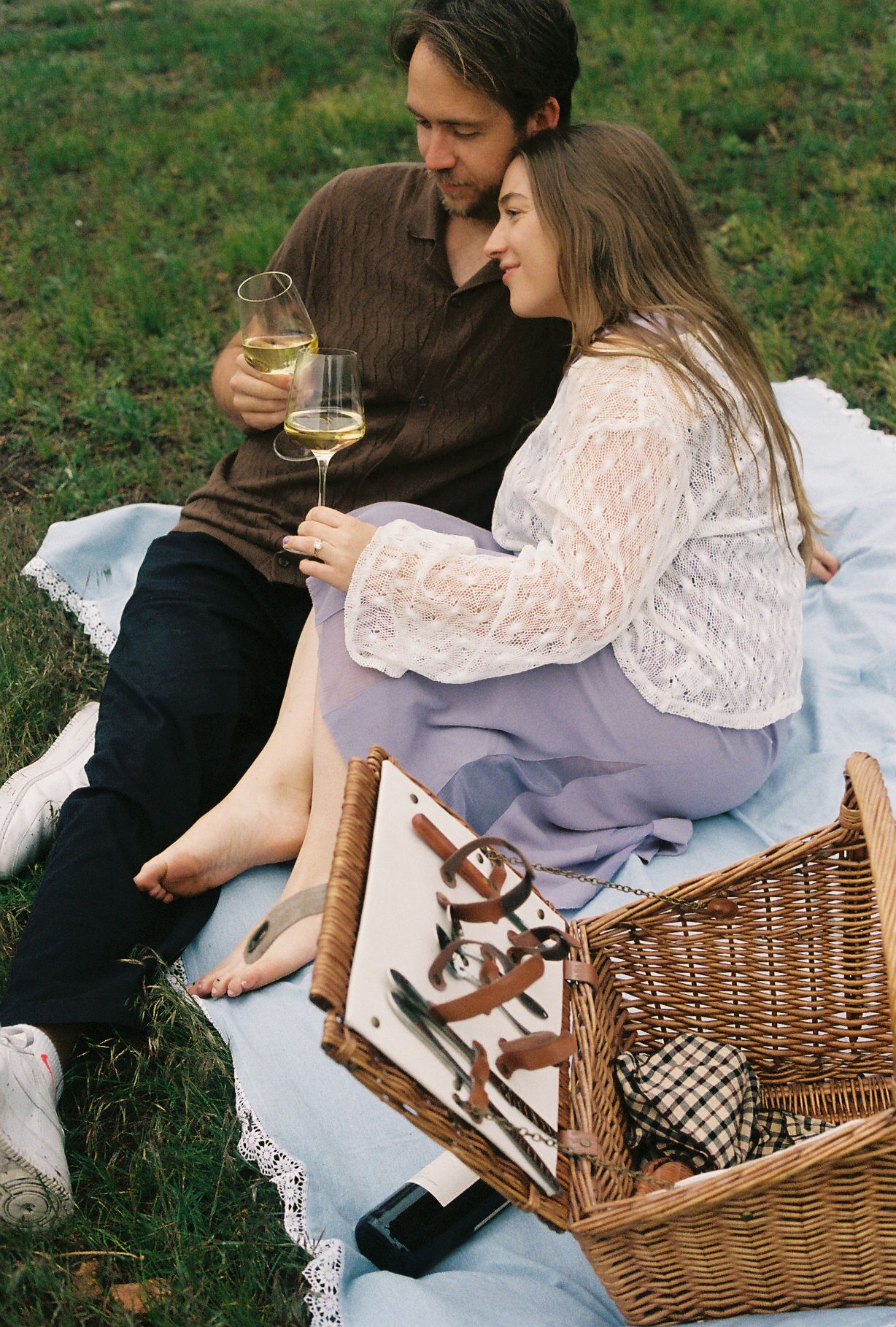 Romantic couple cuddling on a blanket at a picnic with white wine and a wicker basket outdoors.