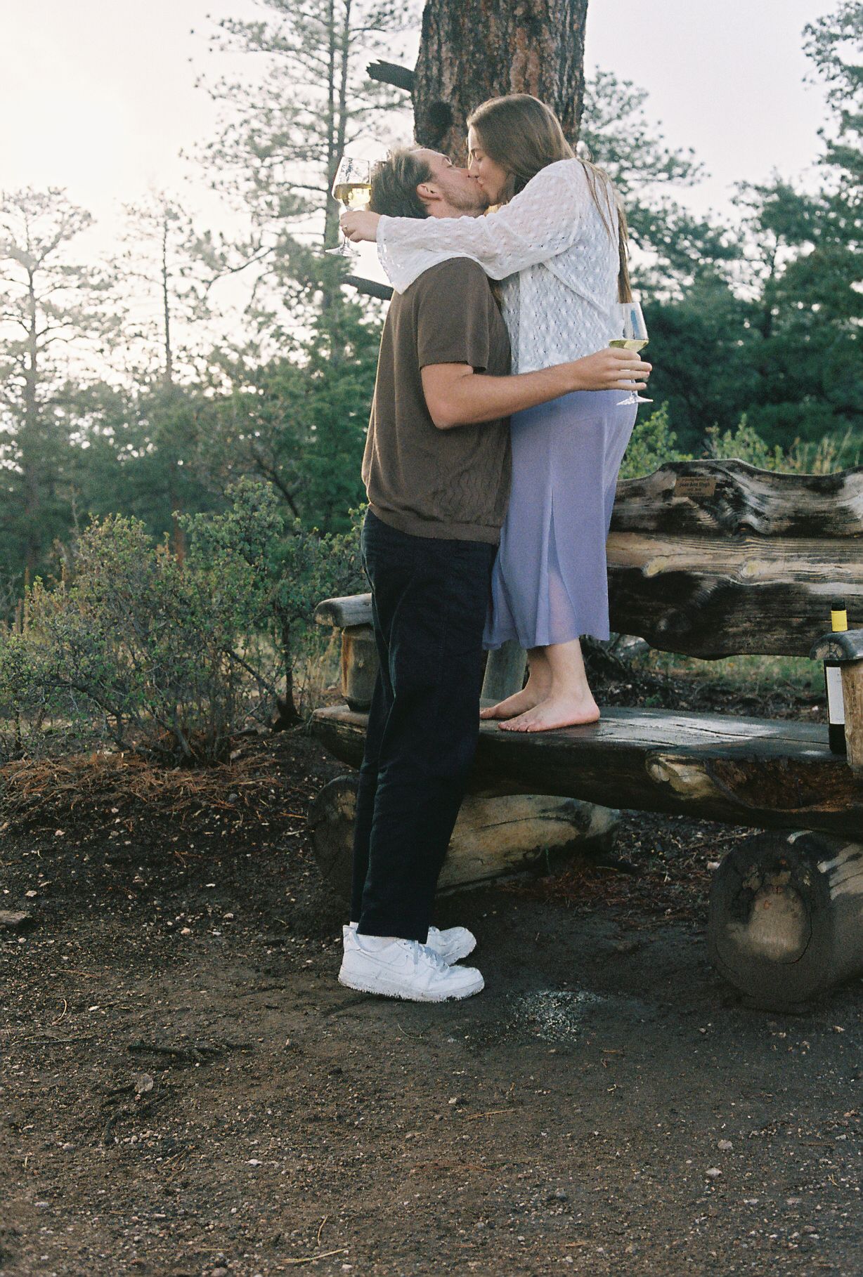 Couple kissing on a rustic log bench in a pine forest while holding glasses of white wine.