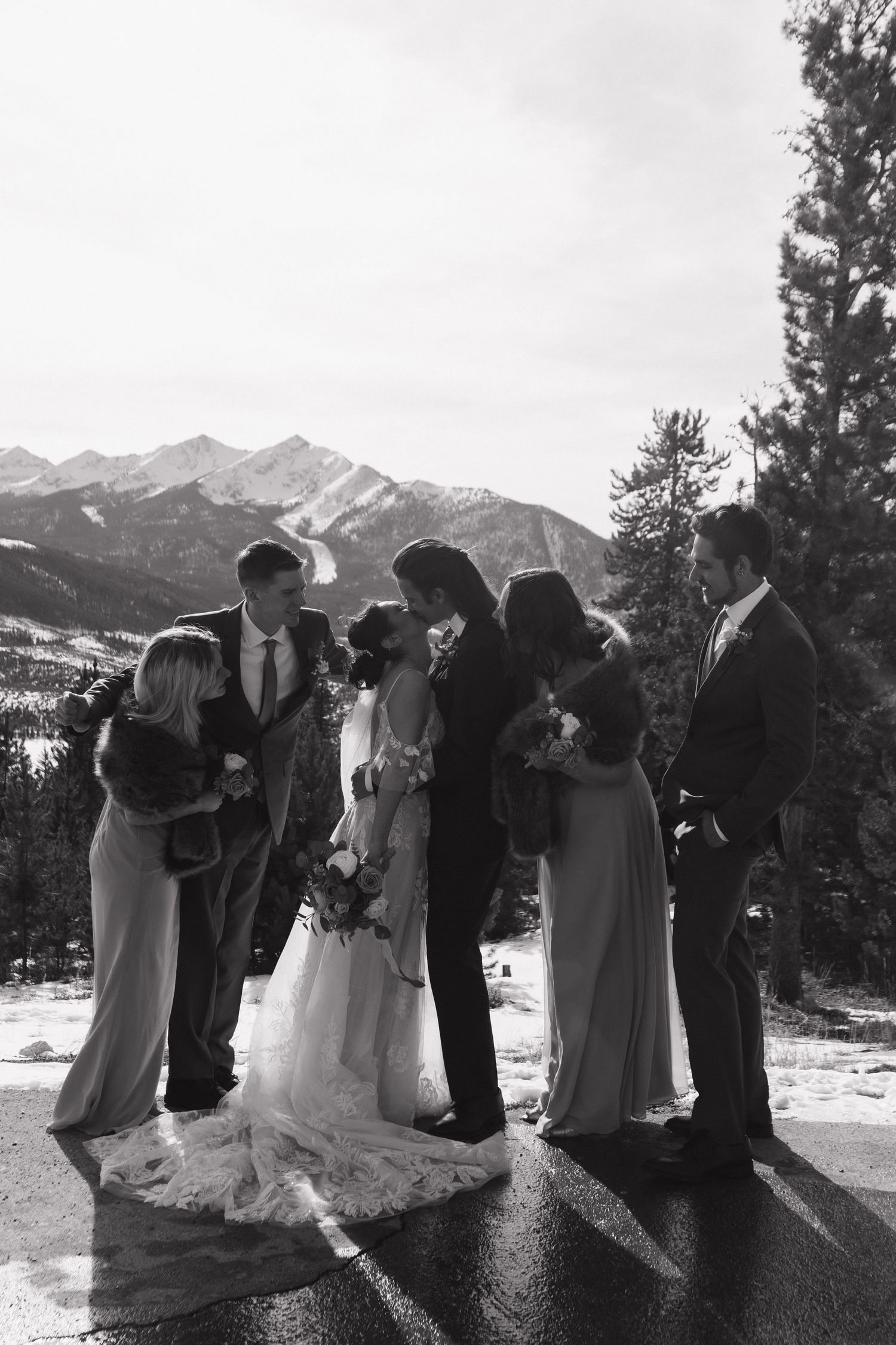 Wedding party gathered on a mountain overlook with snow-capped peaks in the background.