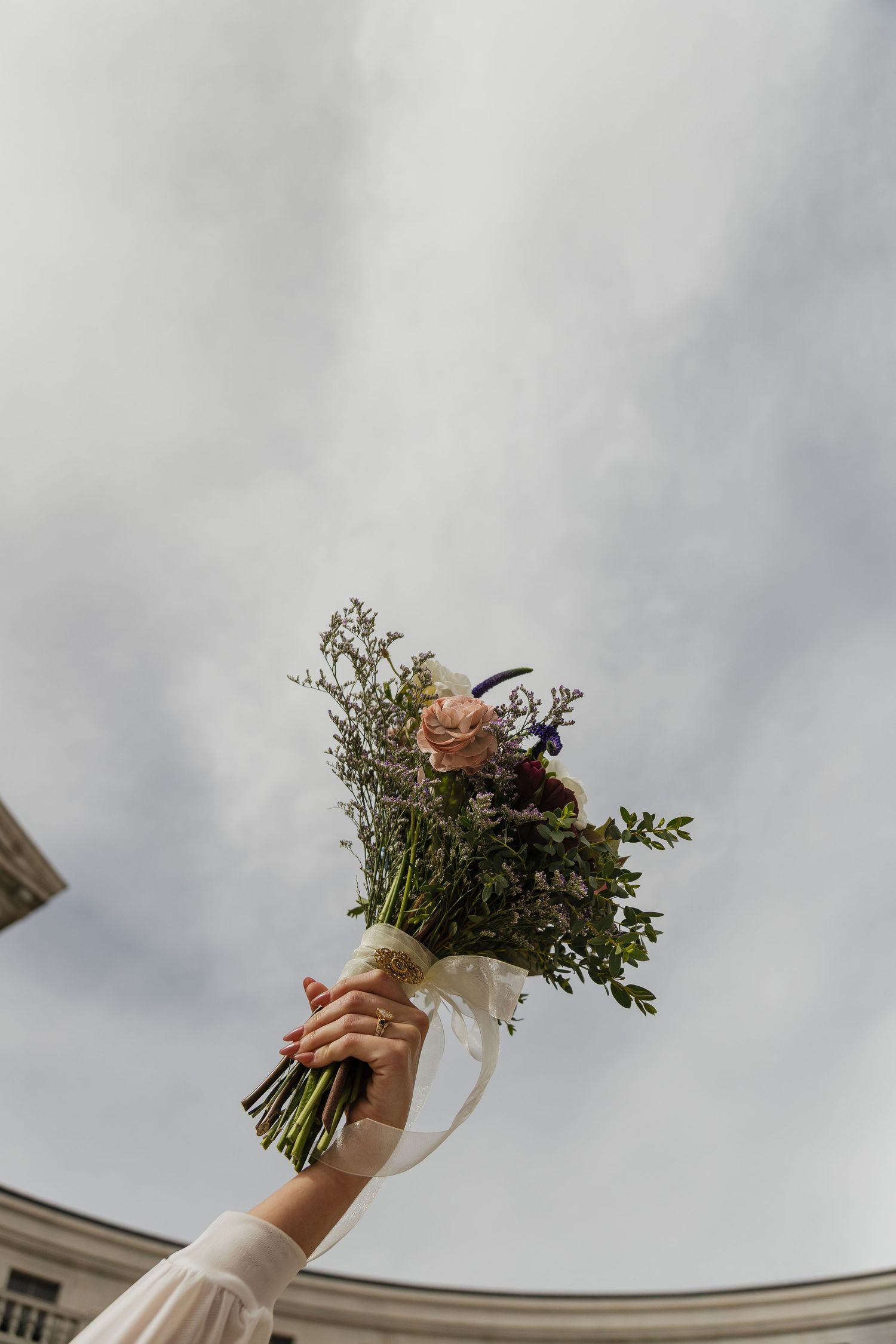 A rustic wedding bouquet with wildflowers is held up against a cloudy sky.