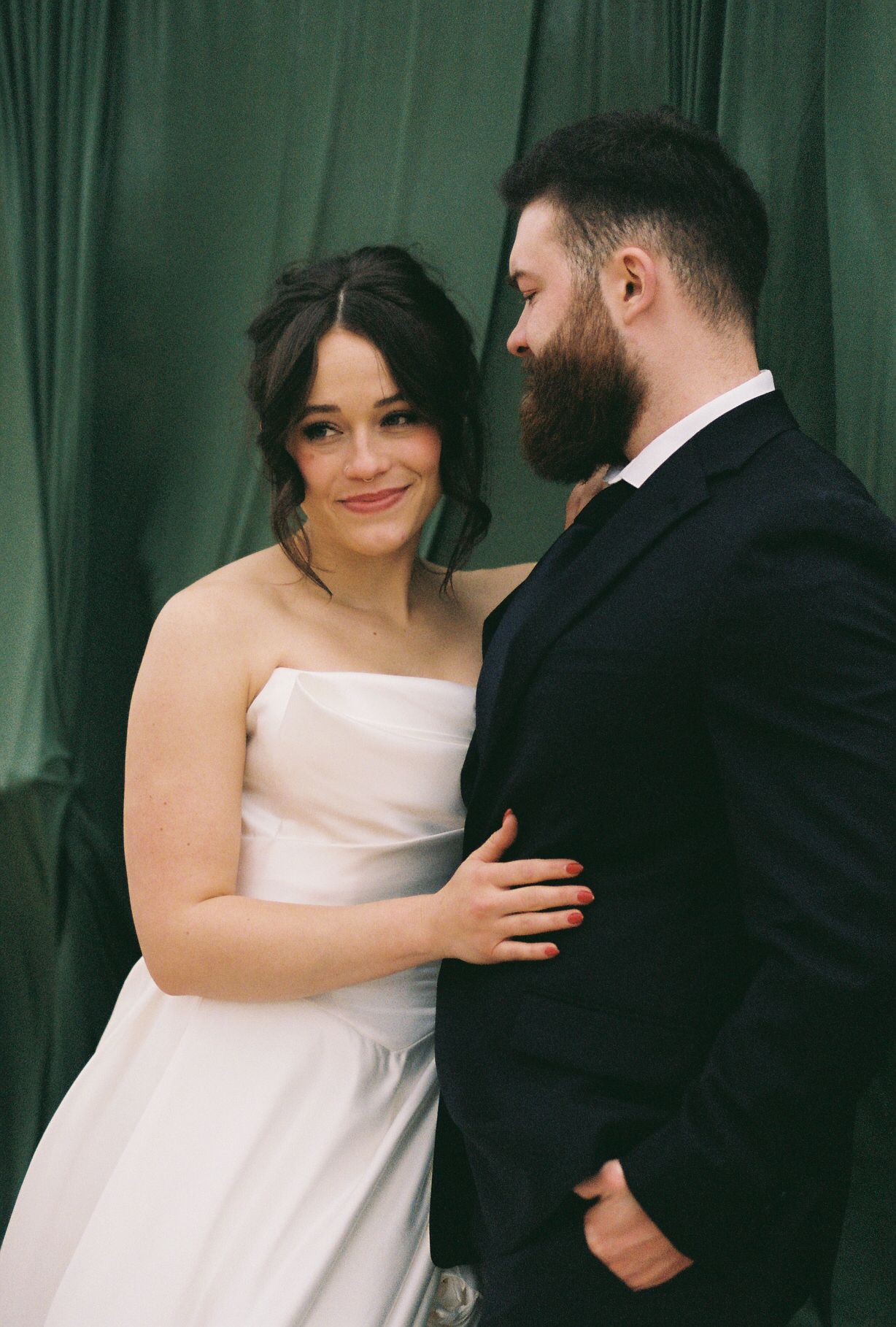 Intimate wedding portrait with couple embracing in front of green curtain backdrop.