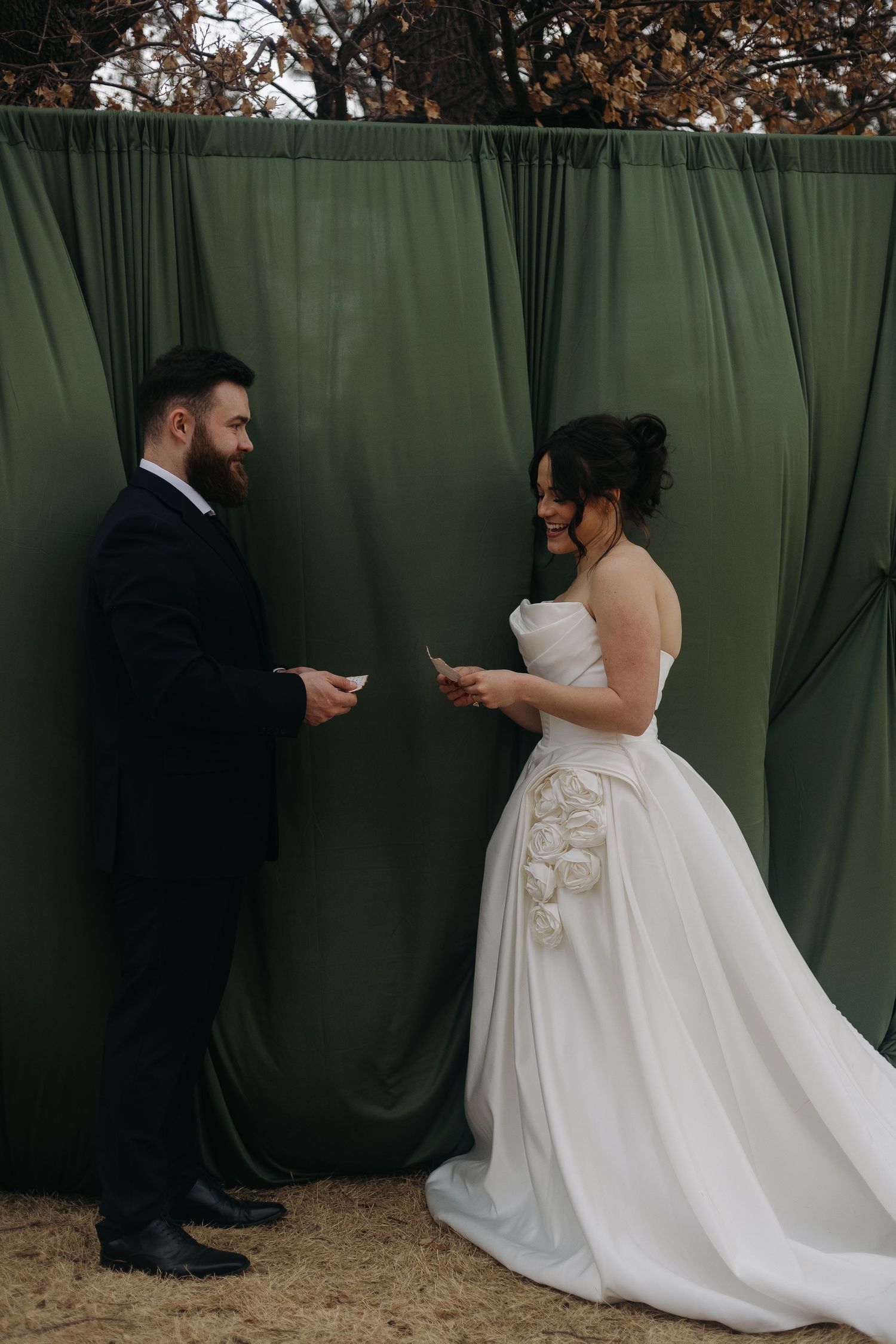 Couple exchanges items during wedding ceremony against green backdrop outdoors.