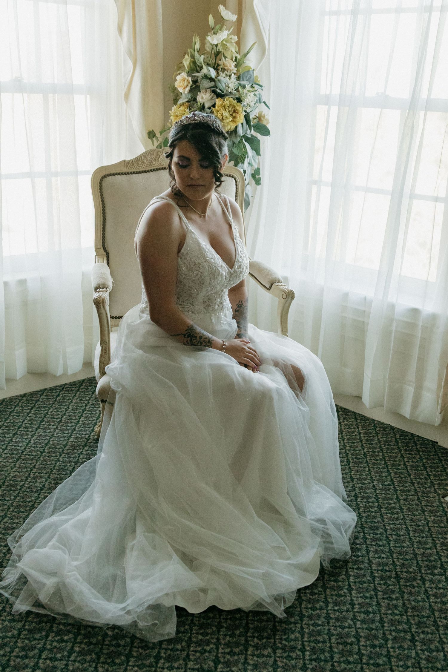 A bride in a flowing white gown sits elegantly on a vintage chair against sheer curtains in a room with patterned carpet.