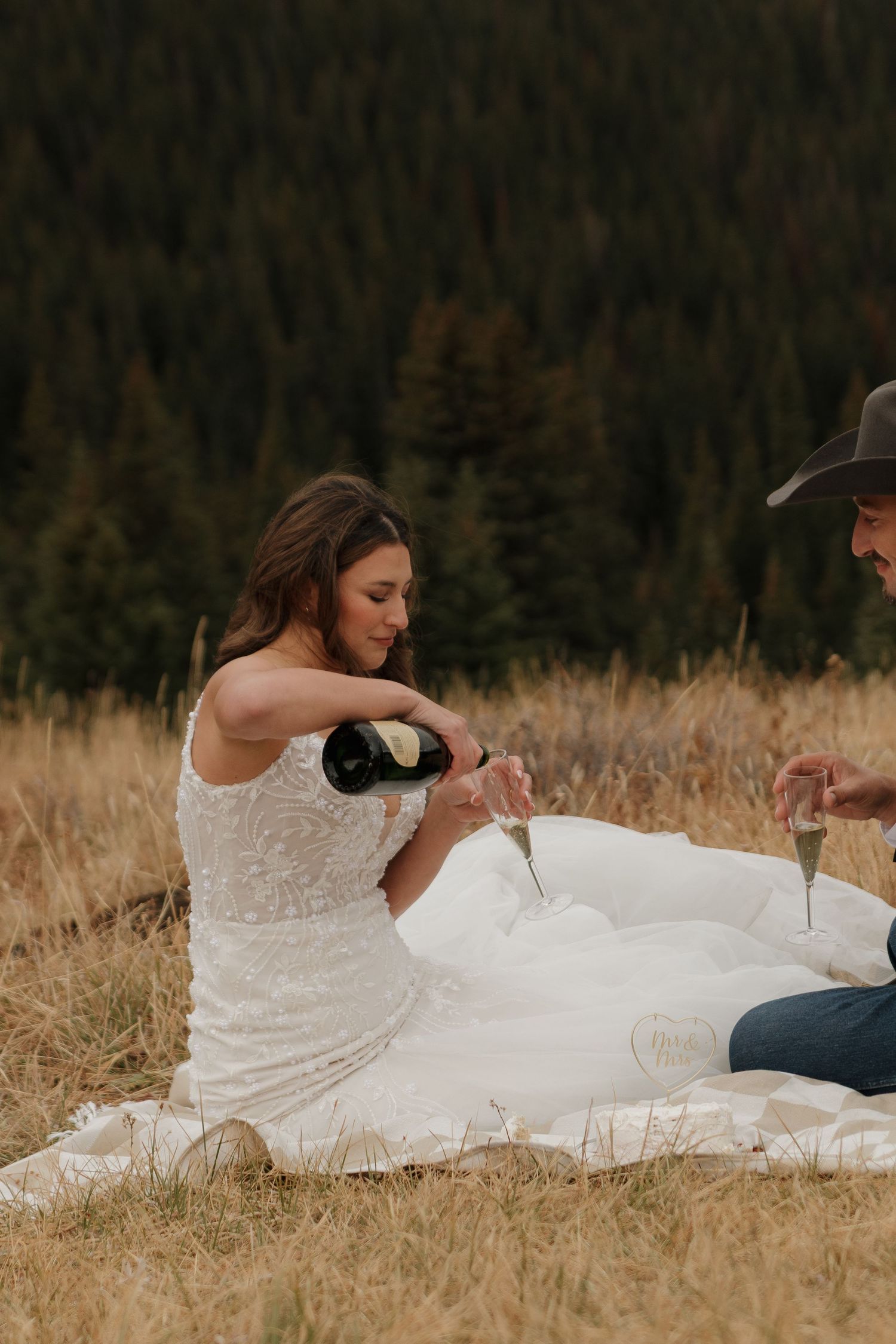 A couple pours champagne while having a picnic on a scenic mountain hillside.