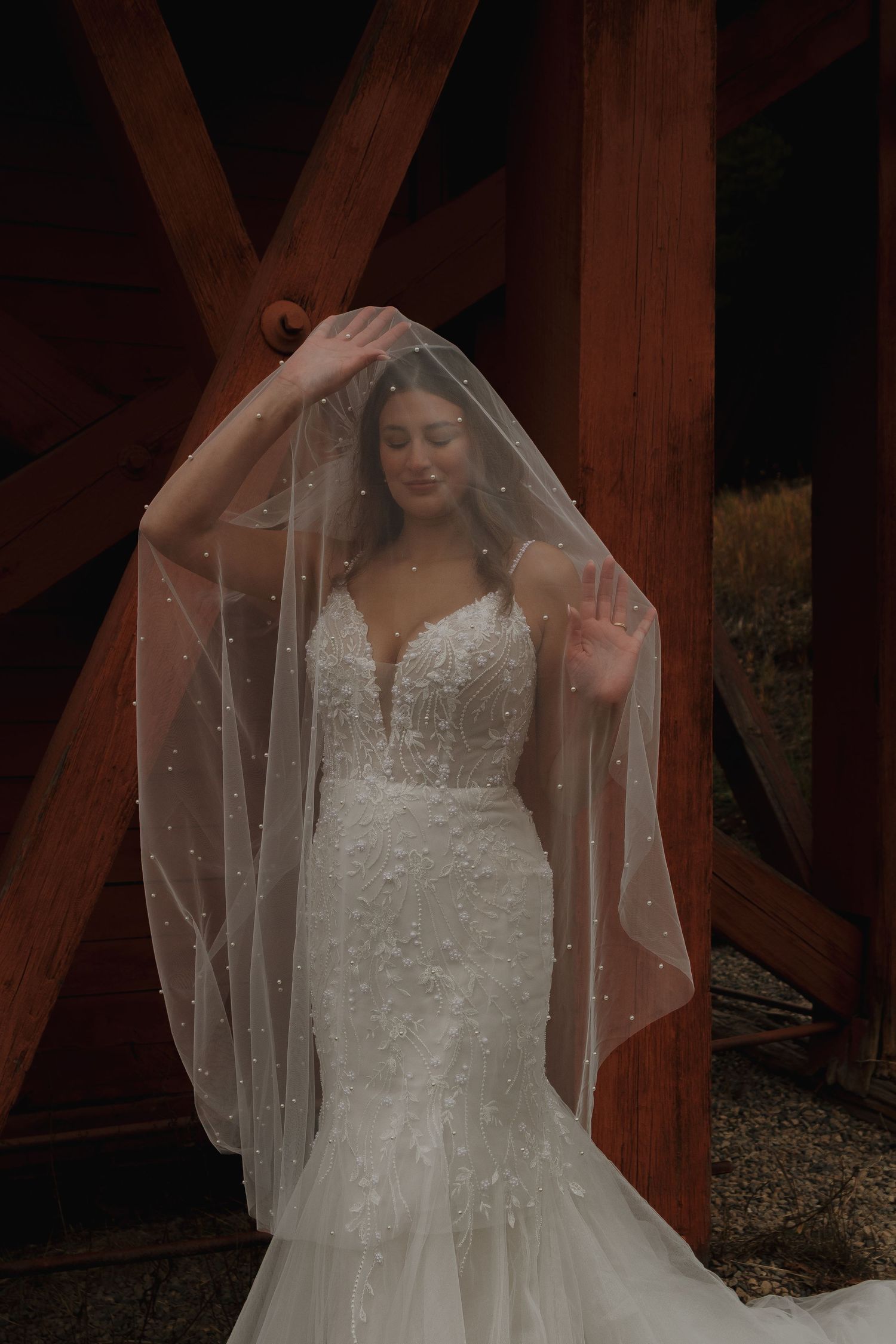 A bride in a lace mermaid wedding dress poses under a delicate veil against wooden beams.