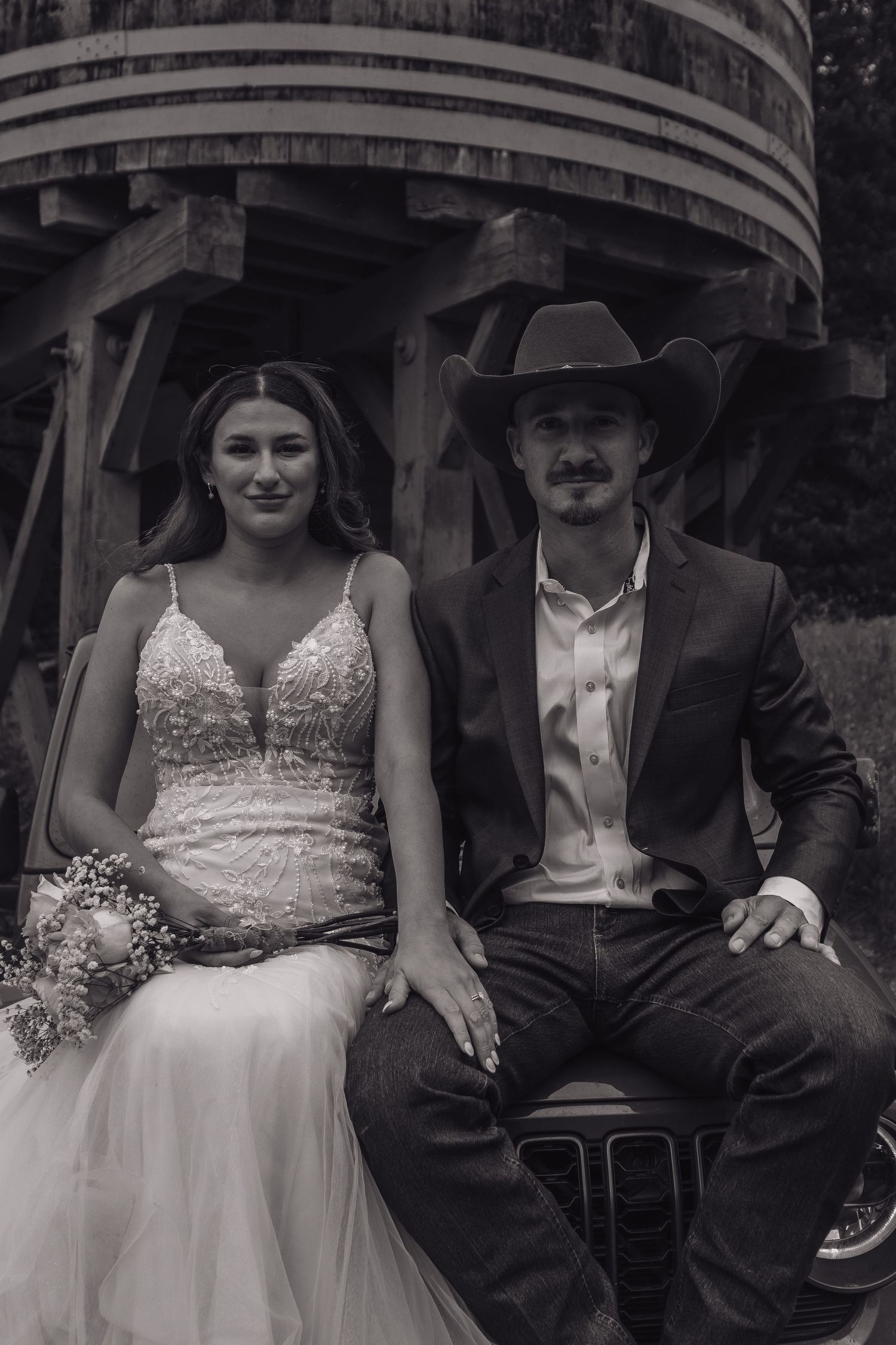 A black and white portrait of a couple in western wedding attire seated beneath wooden architecture.