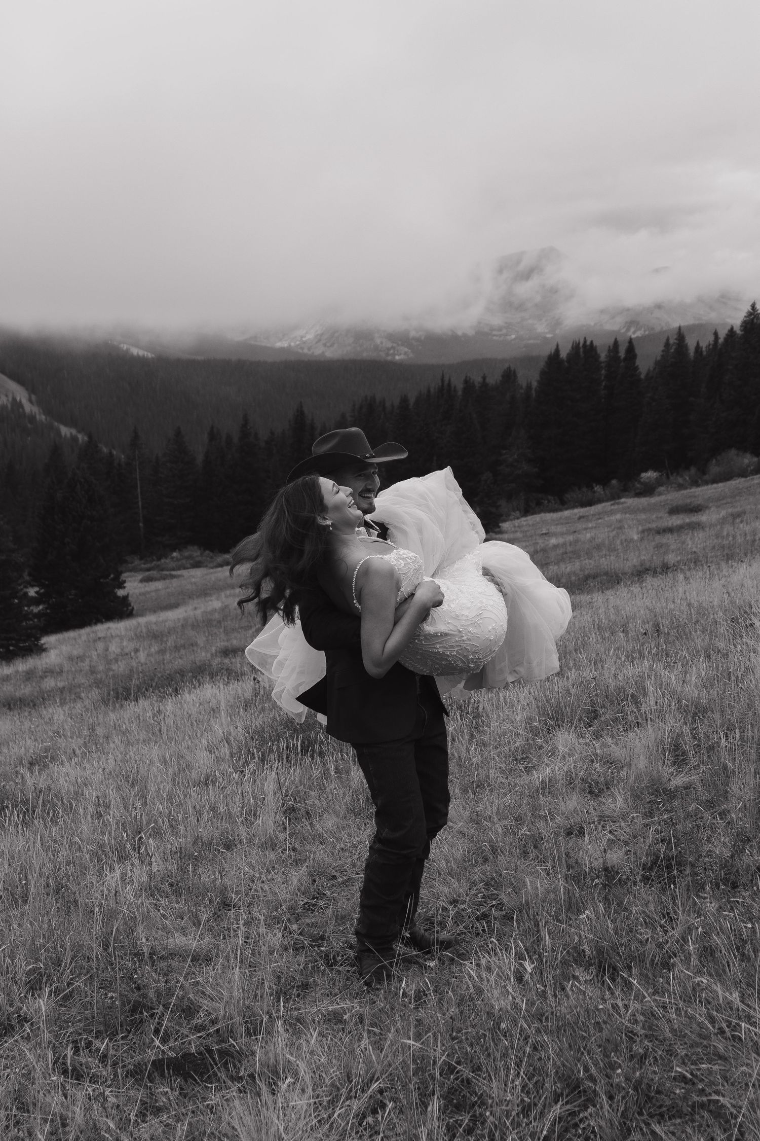 A black and white wedding photo captures a romantic dip in a mountain meadow.