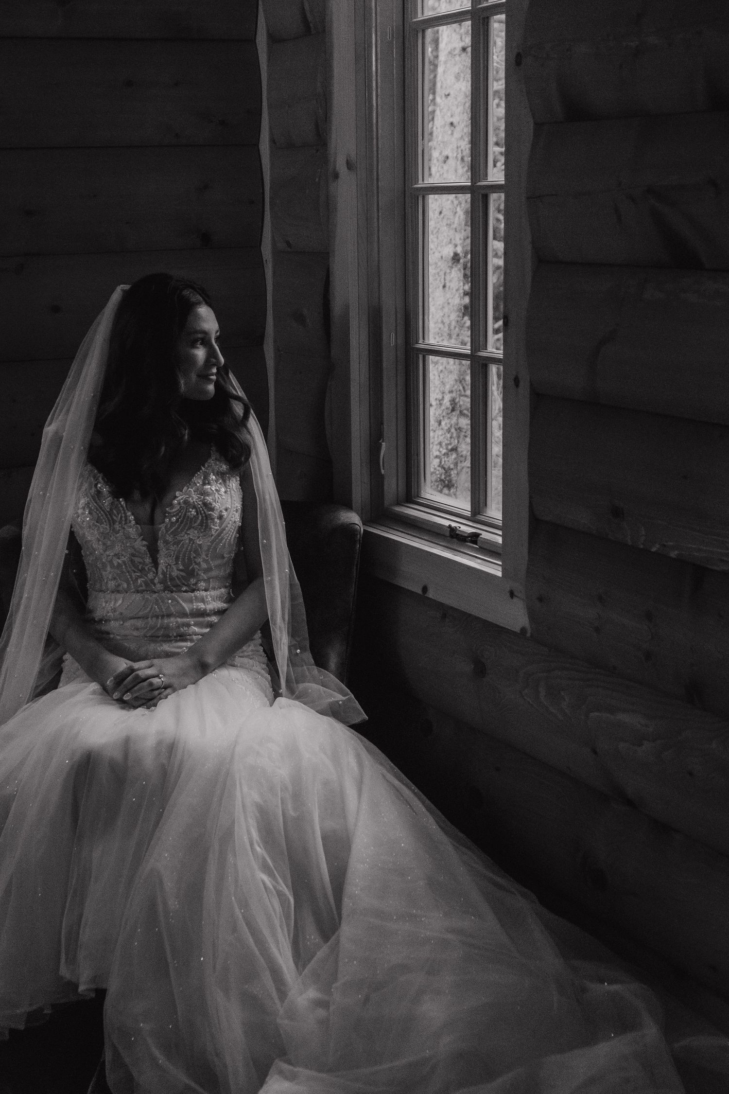 Dramatic black and white photo of a bride sitting by a window in a rustic cabin.
