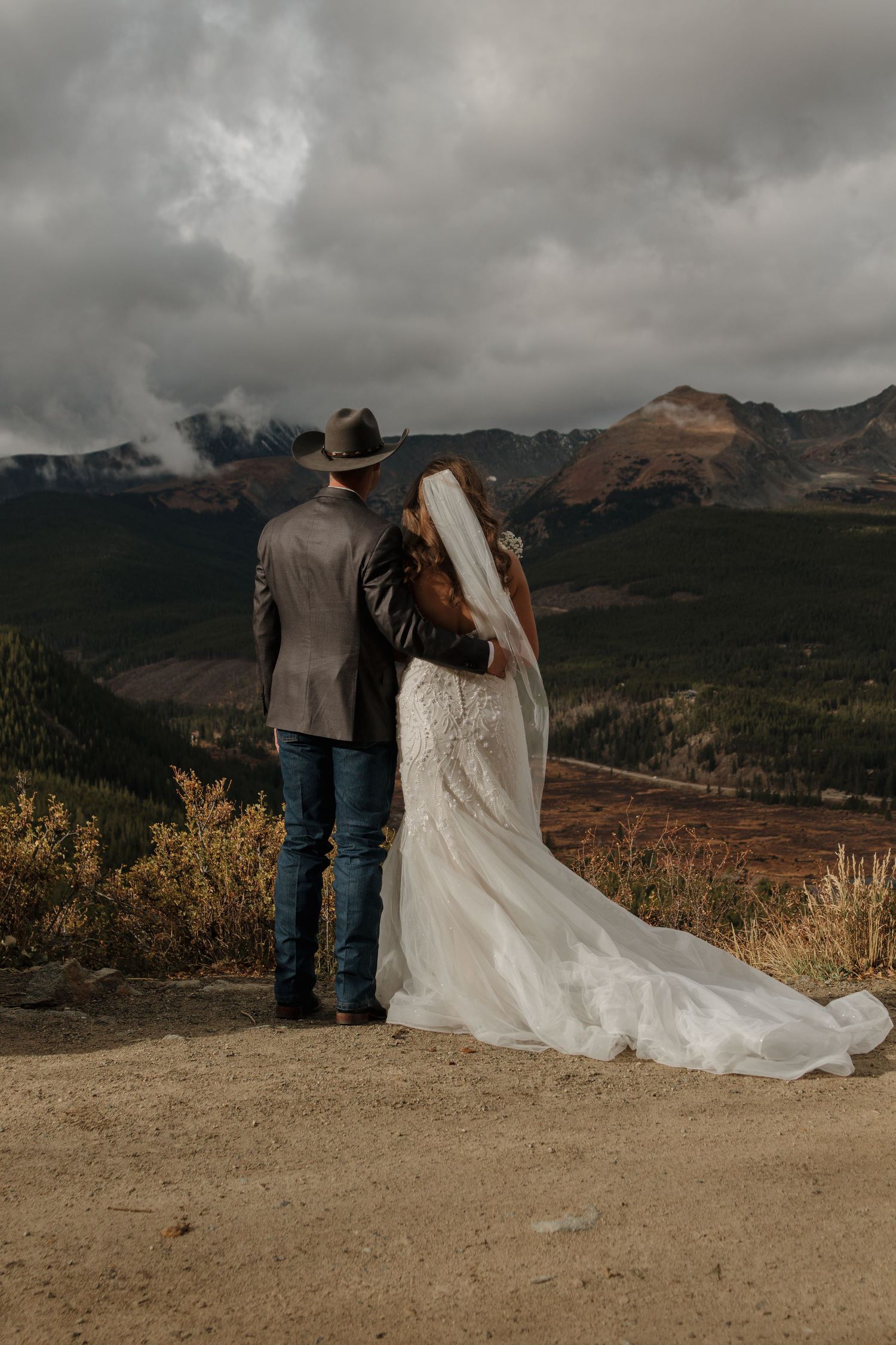 A couple in wedding attire stands on a mountain overlook with dramatic cloudy skies in the background.