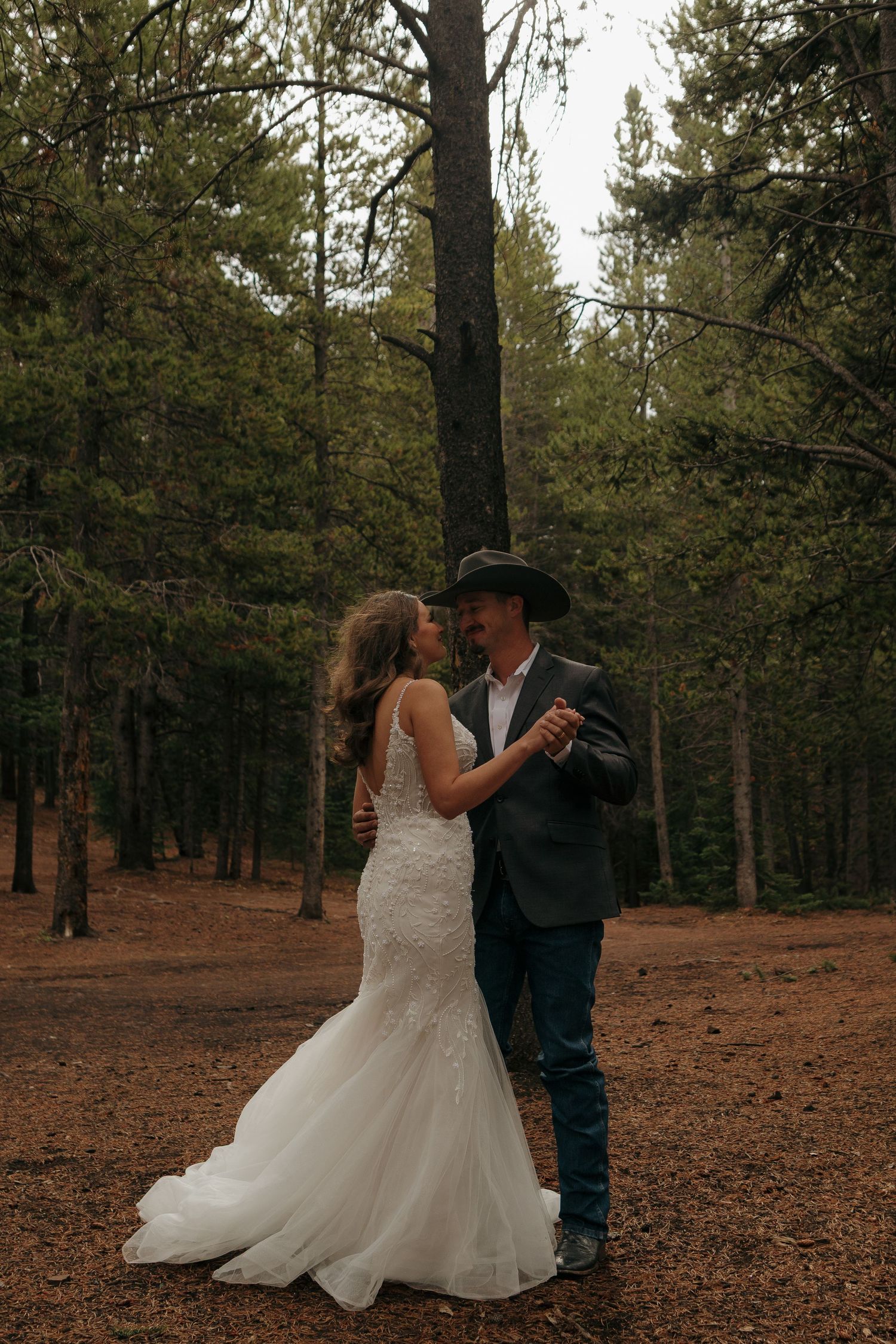 Newlyweds share an intimate dance among pine trees on a forest floor covered in fallen leaves.