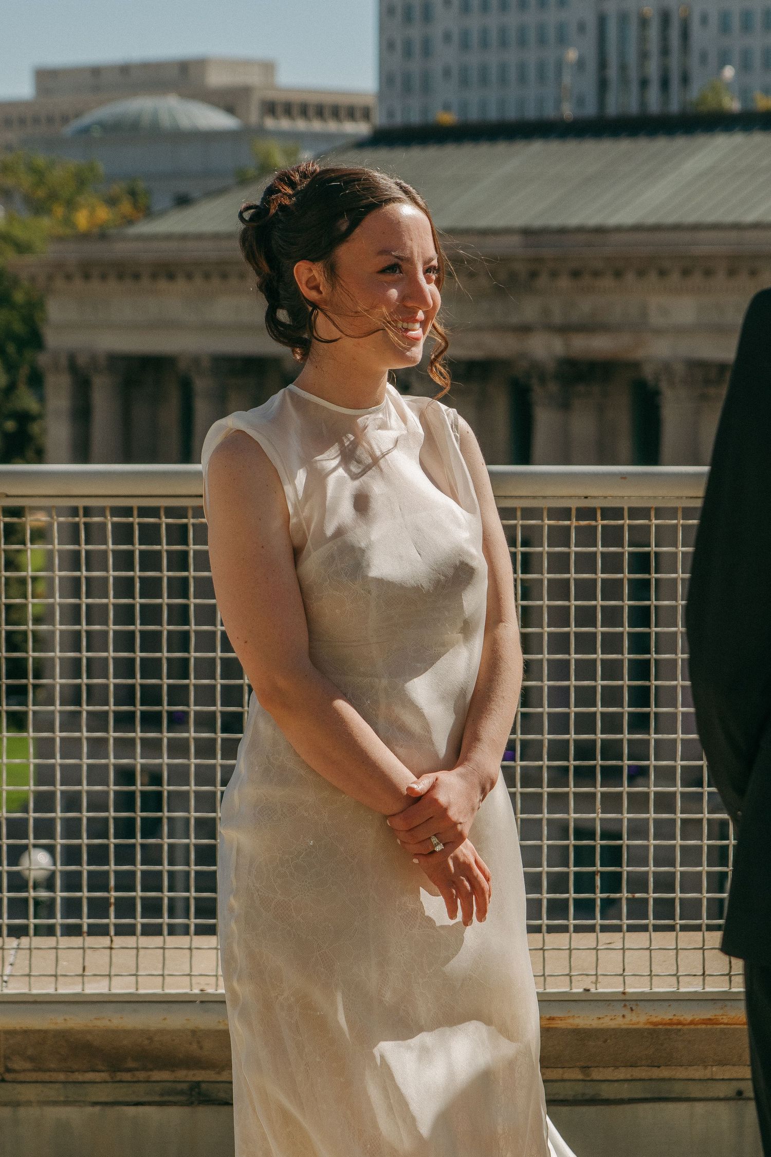 Bride in white dress standing on outdoor terrace with city architecture in background.