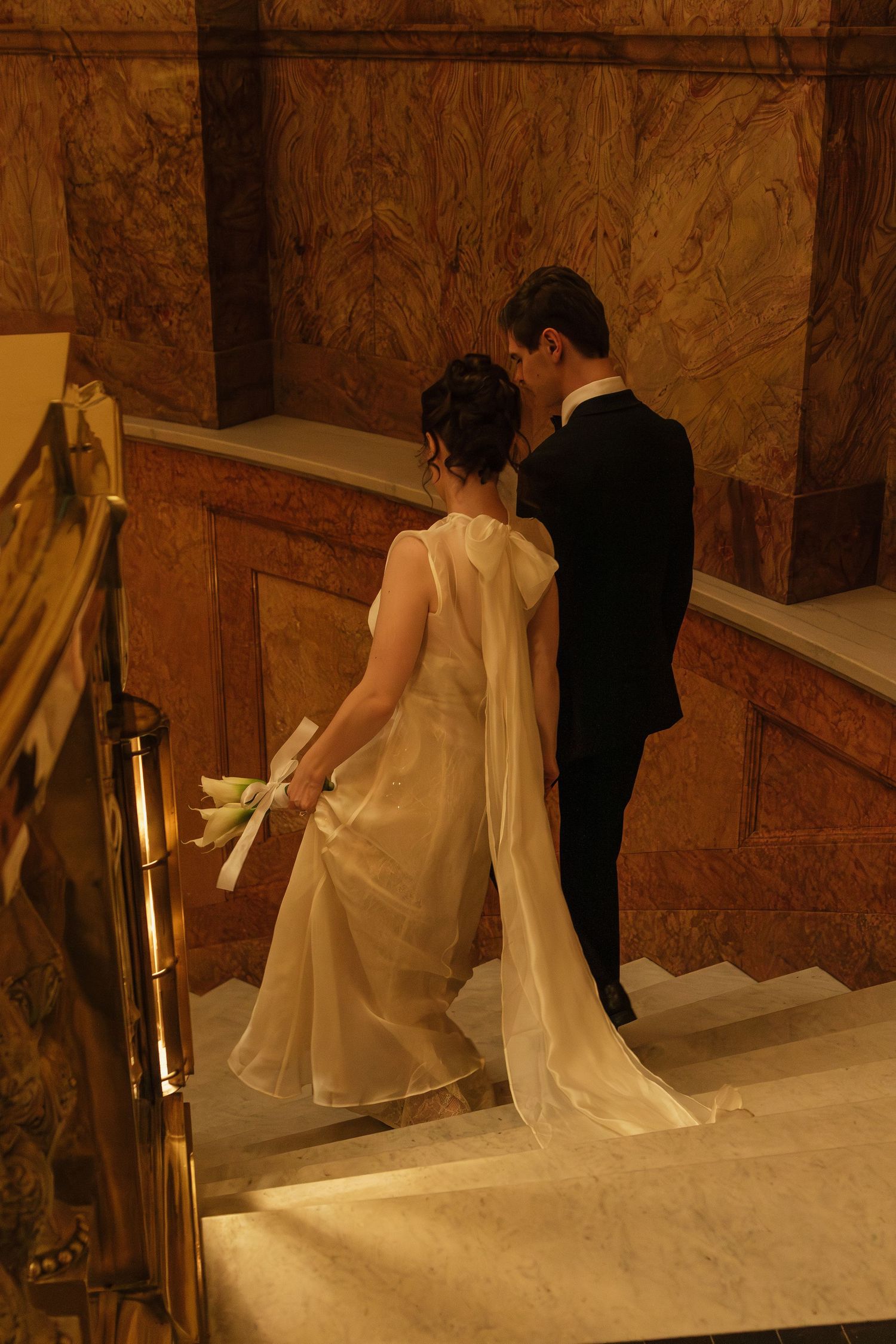 Couple in wedding attire walking down marble stairs in warm ambient lighting.