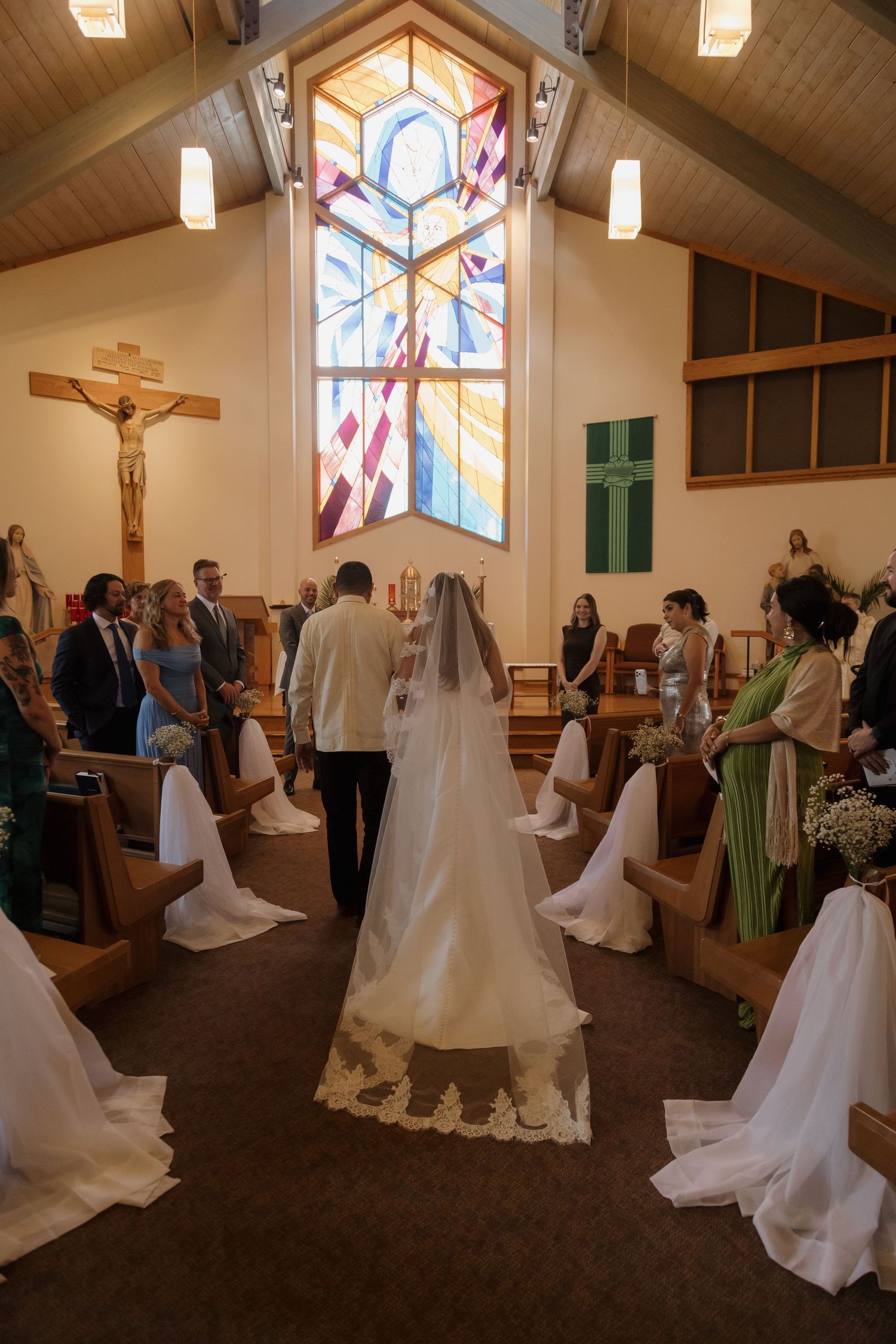 Bride walking down the aisle in a Catholic church with stunning stained glass window during wedding ceremony.