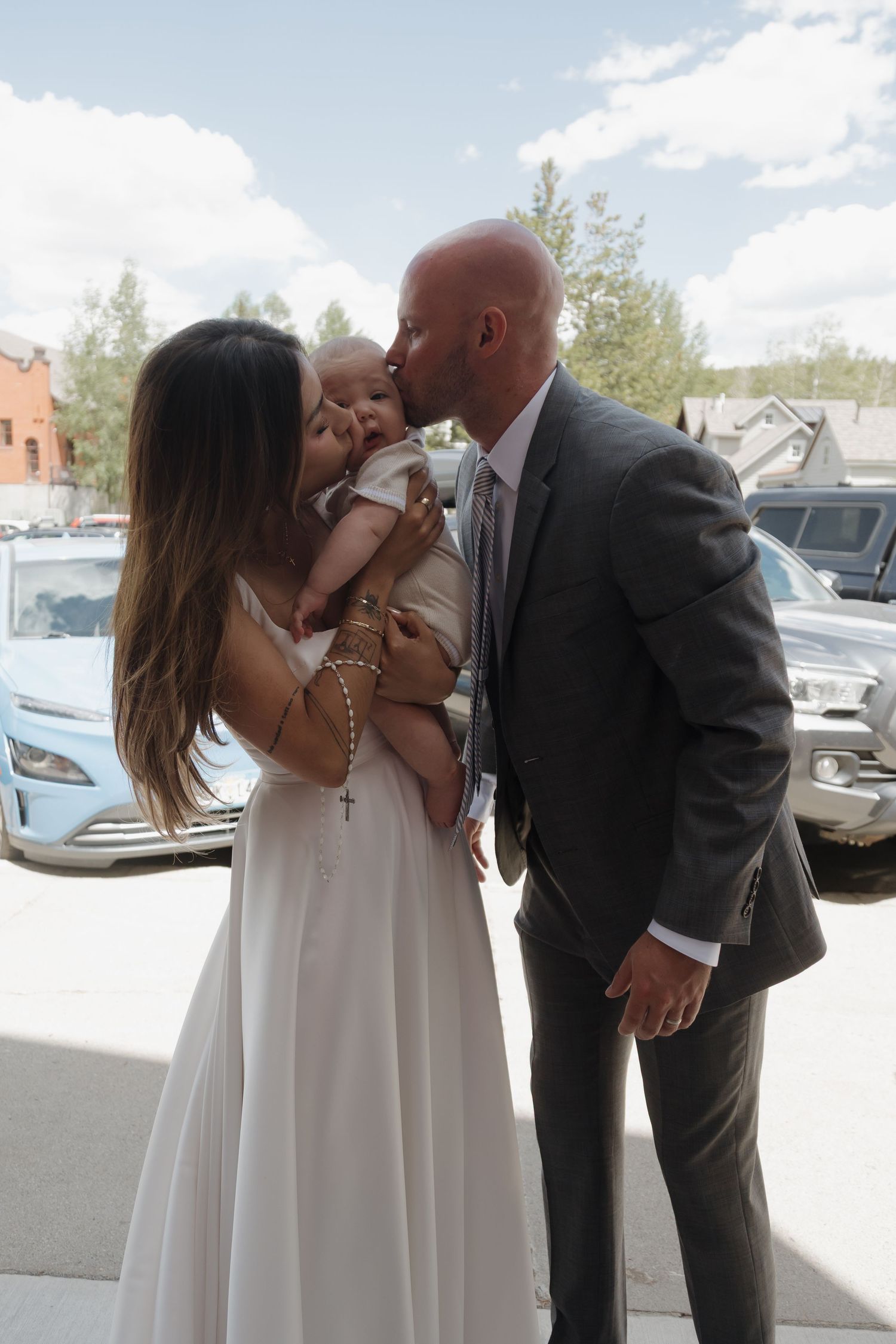 A bald groom in a gray suit kisses a baby held by his bride outside a church.