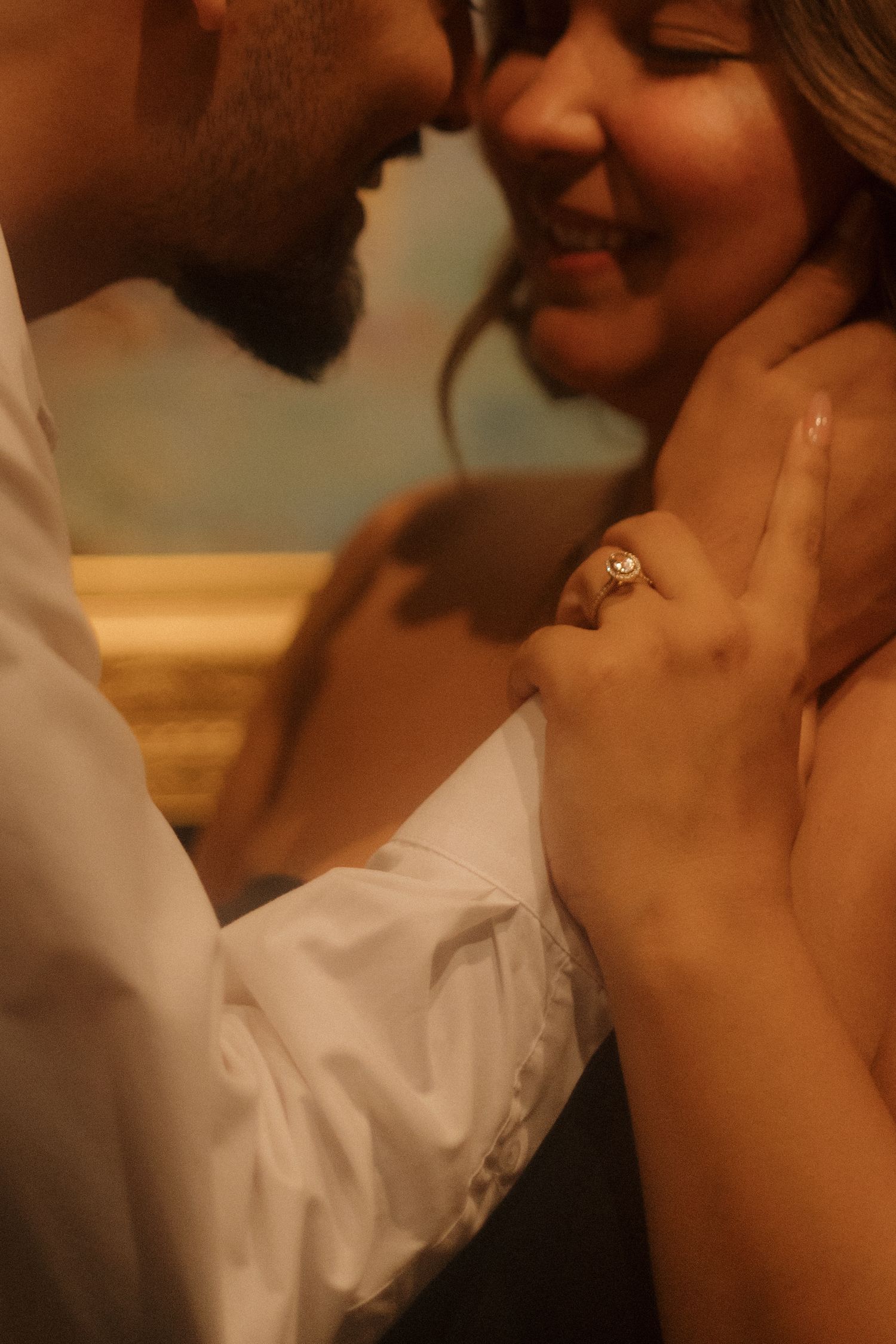 Close-up of smiling couple nearly kissing, woman displaying a sparkling oval engagement ring on her hand.