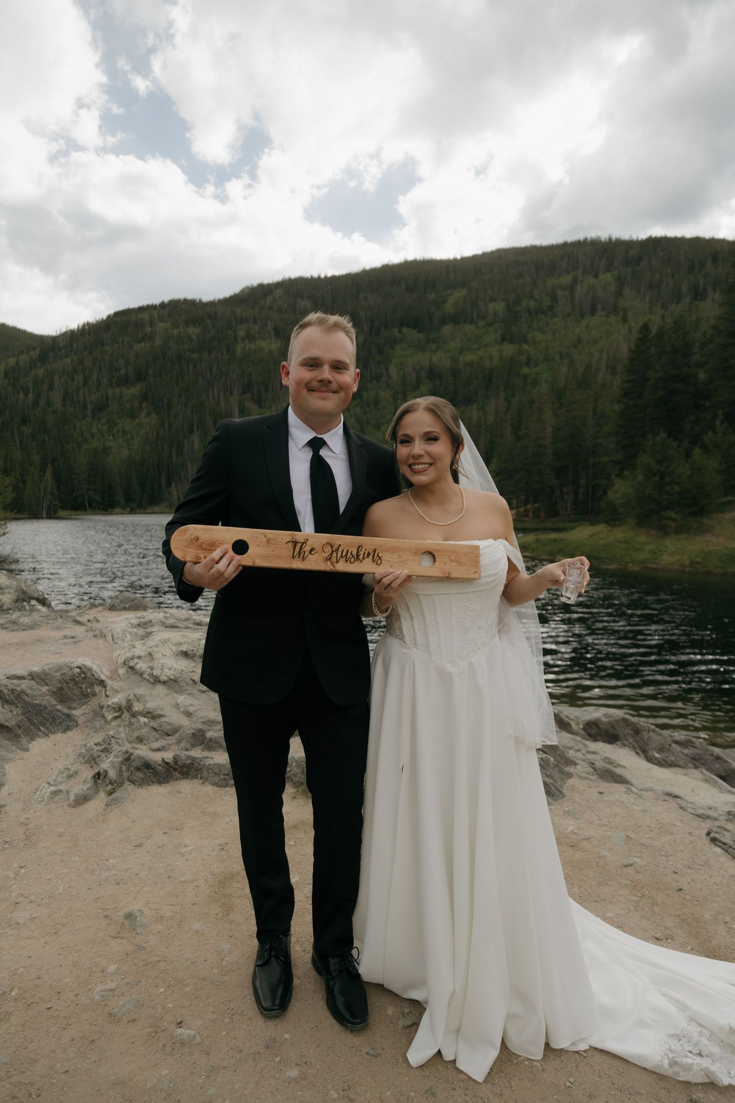 Bride and groom pose with personalized wooden sign by mountain lake on their wedding day.
