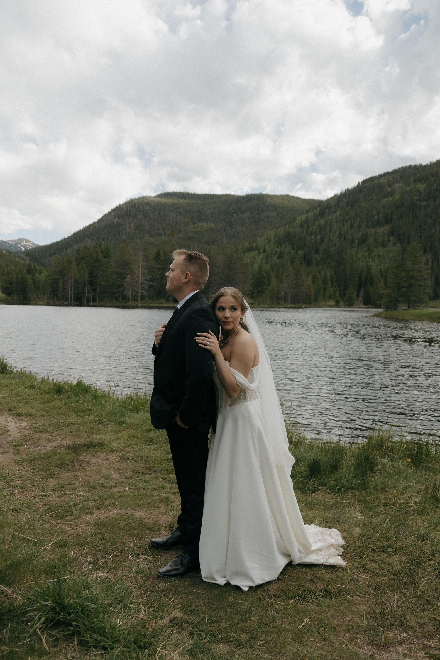 Bride and groom pose back-to-back by a serene mountain lake on their wedding day.