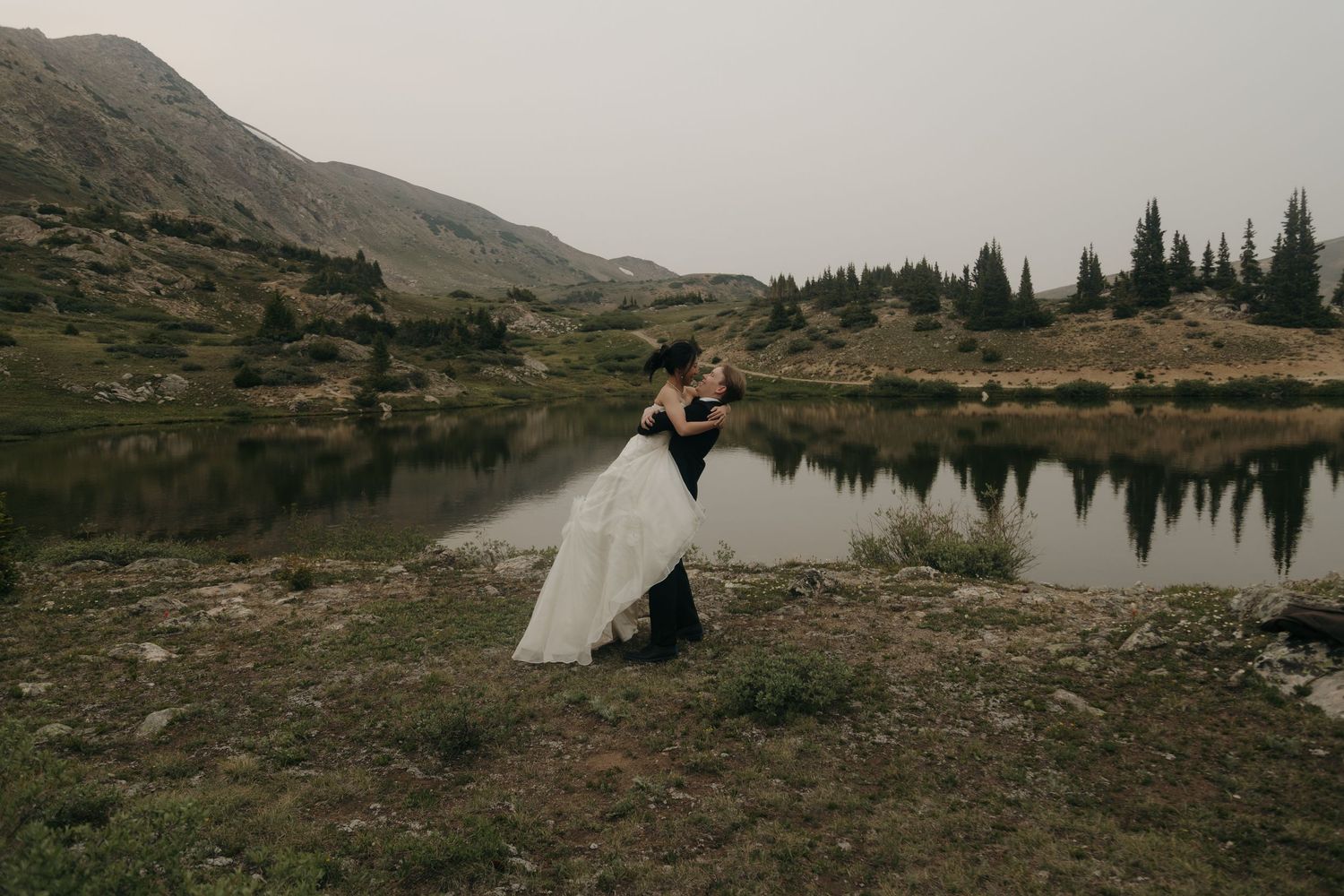 Groom lifts bride in wedding dress by a serene alpine lake surrounded by mountains and pine trees.