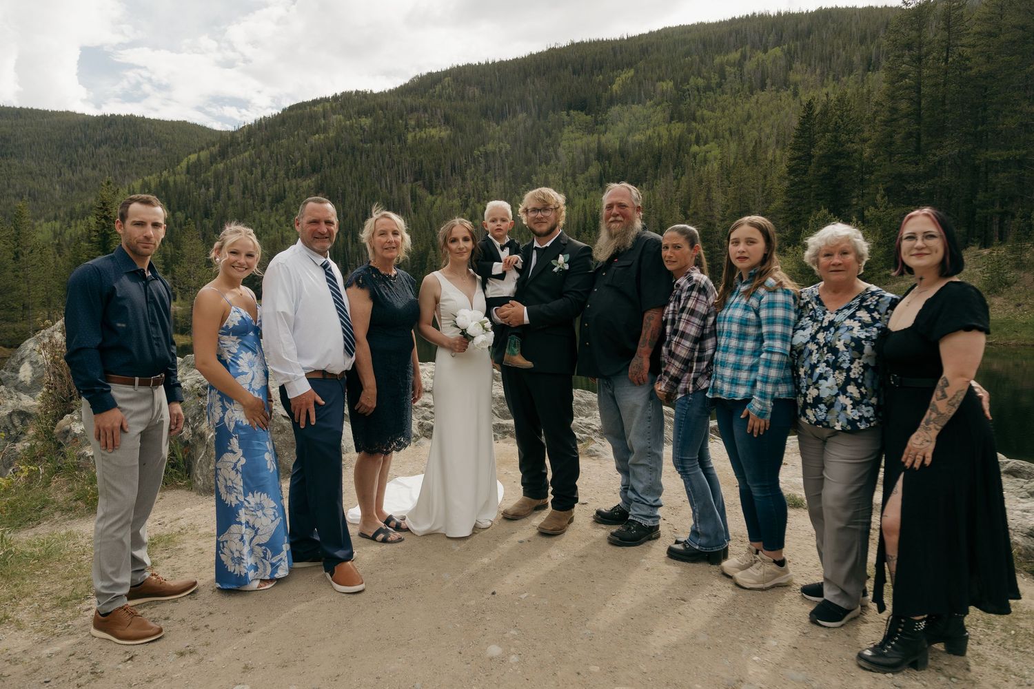 Wedding party group photo with bride and groom holding baby outdoors in mountain setting.